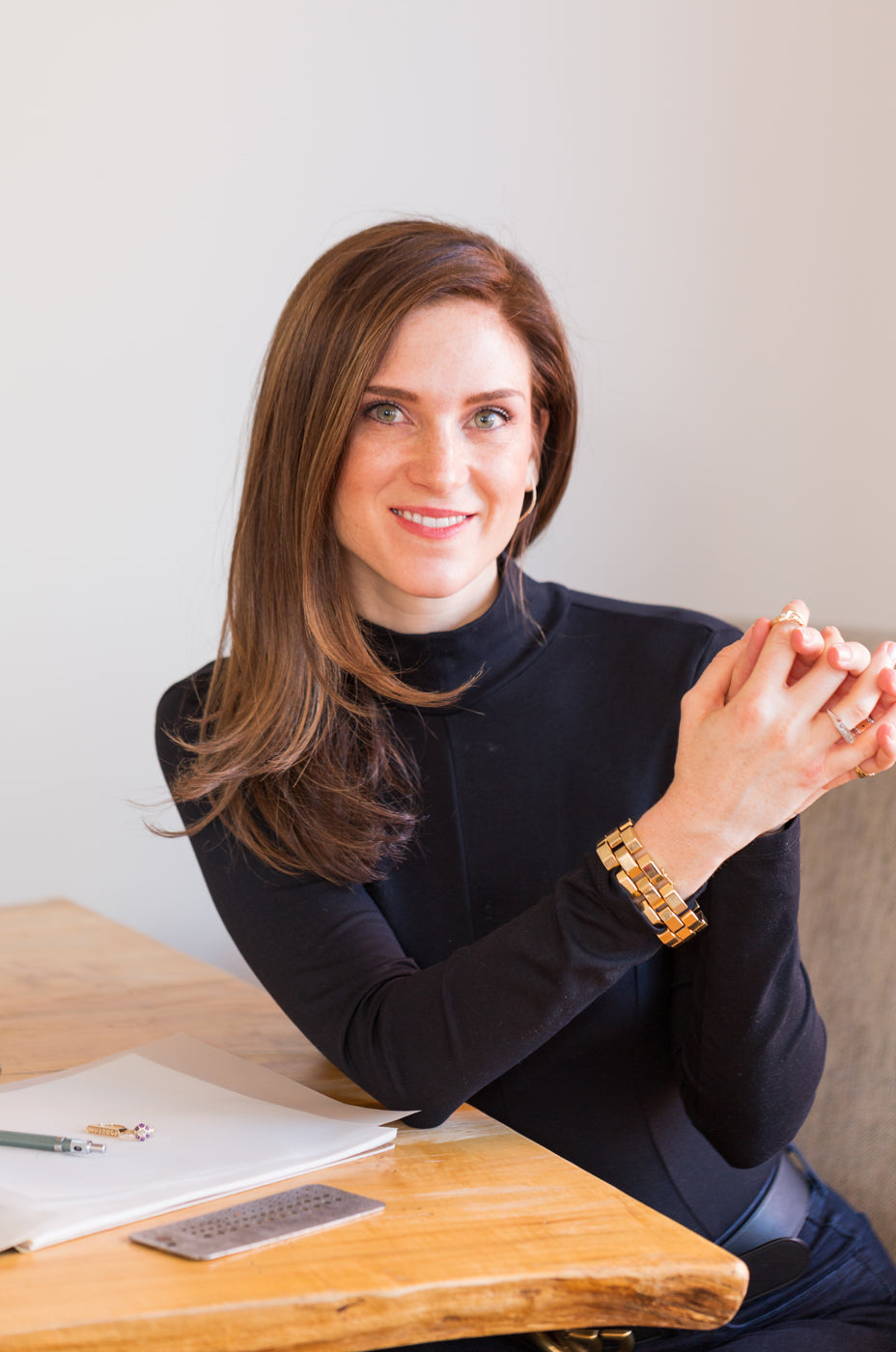 An image of a smiling woman with long brown hair seated at a wooden table against a white background