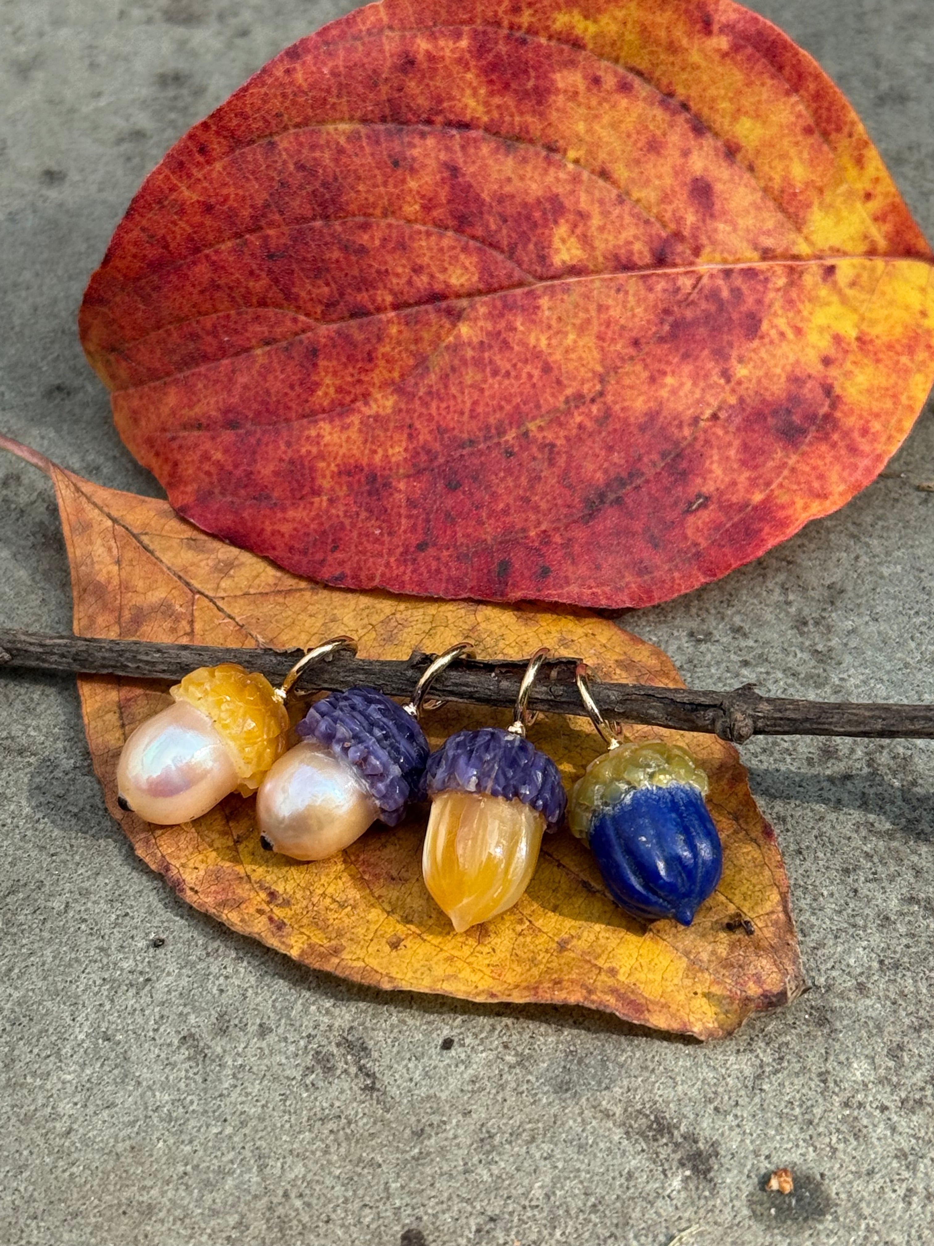 Colorful acorn charms in pearls, golden hues, and blues on a leaf with a red leaf on a concrete surface