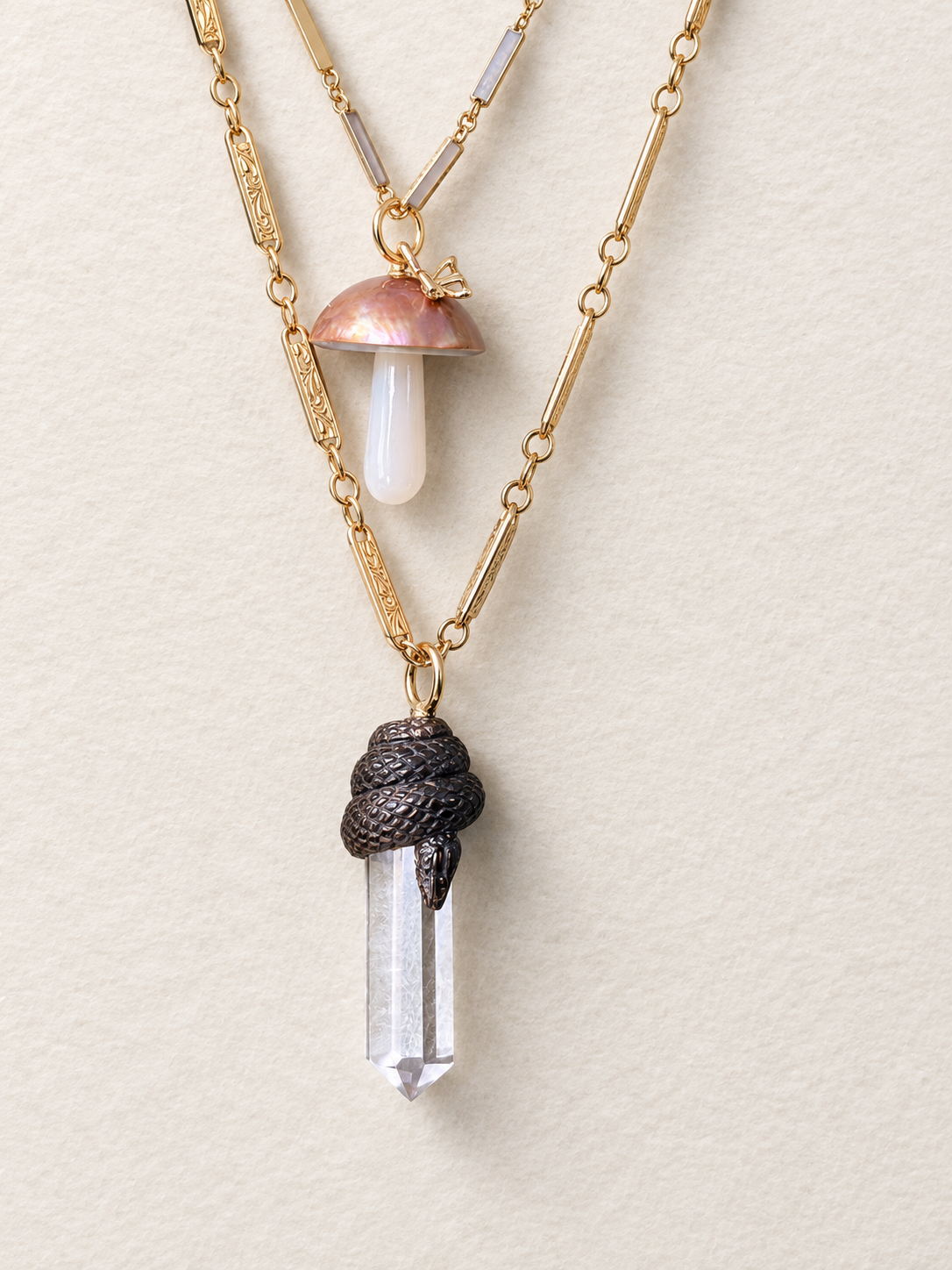Layered 14K yellow gold necklaces featuring a black carved snake coiled around a clear chalcedony crystal point and a mother-of-pearl mushroom pendant with an iridescent shell cap.