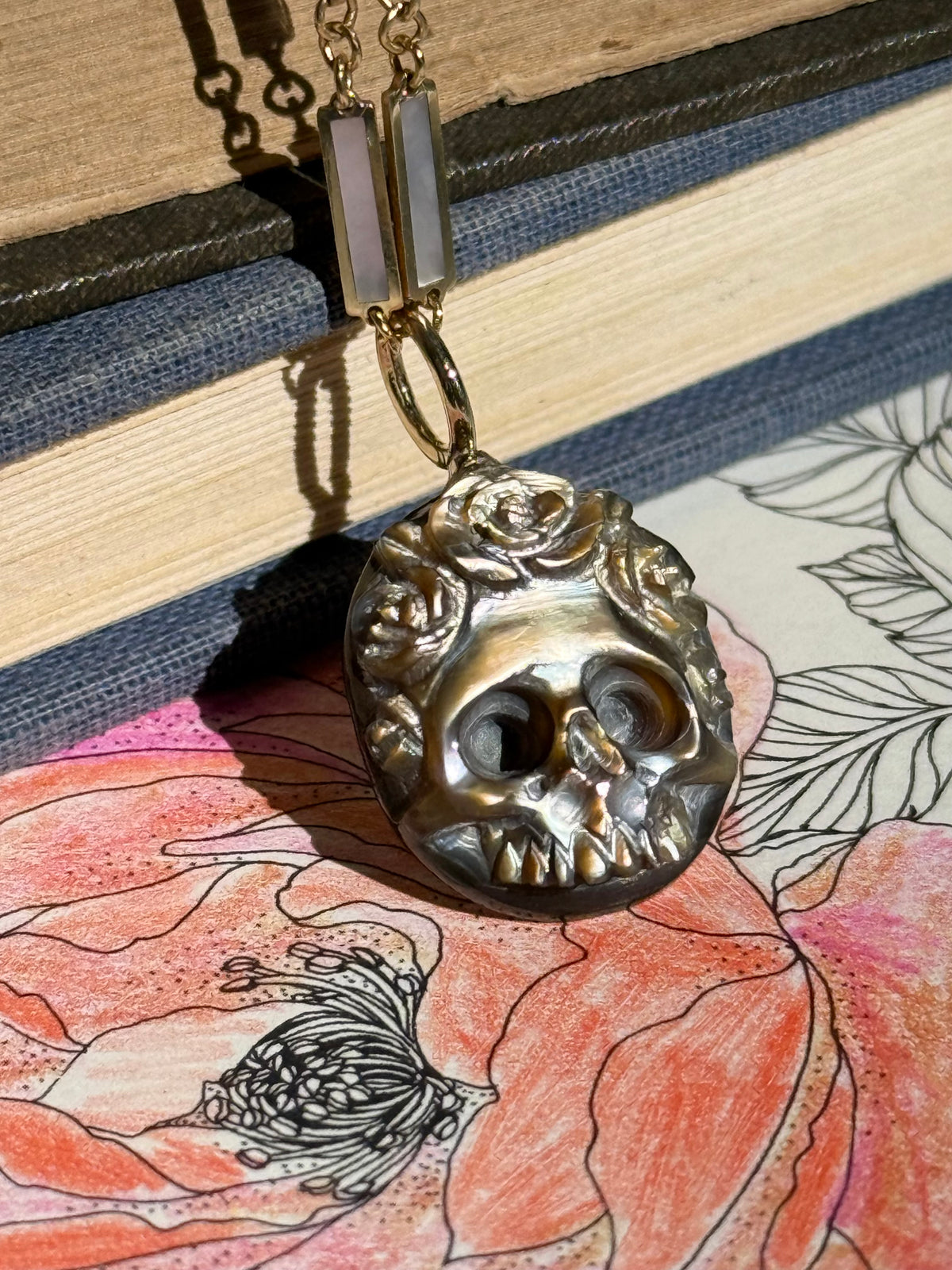 A hand carved brown mother of pearl skull charm with floral details around the crown and a gold round bail leaning against some antique books with a colored pencil flower in the foreground.