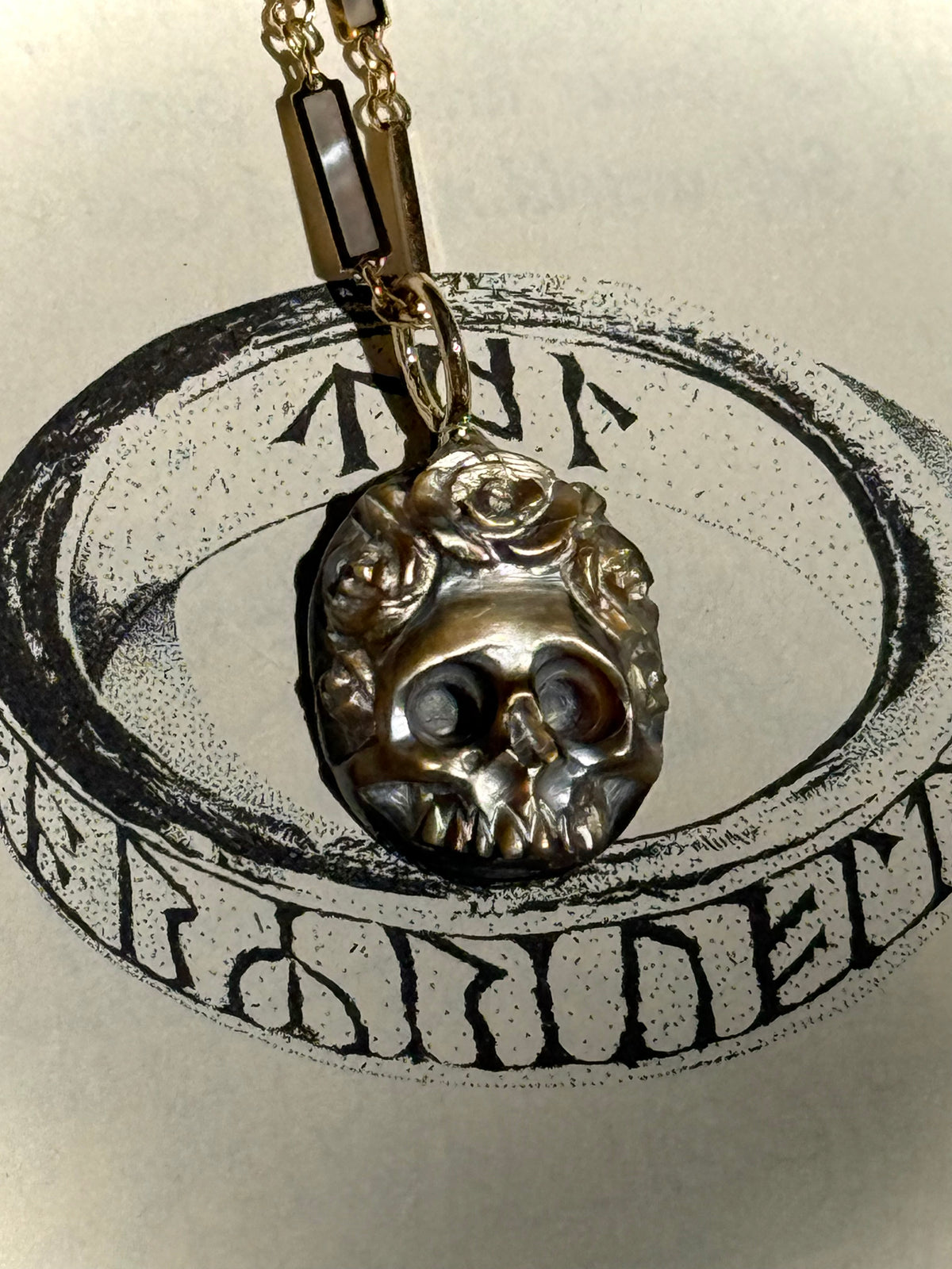 A hand carved brown mother of pearl flower skull charm with a gold bail resting atop the page of a book which has a black illustrated ring with ancient runes engraved around it.