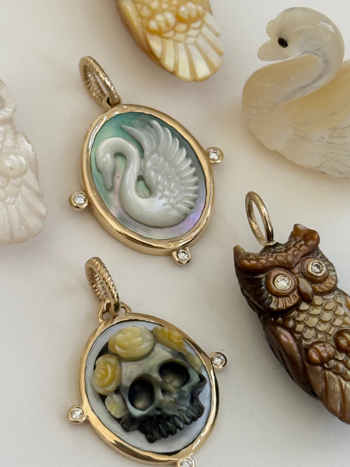 Two cameo style oval pendant charms with gold-tone borders displayed on a light surface. The top pendant features a carved white swan on a pale green iridescent background, and the bottom pendant shows a black-and-white skull with yellow flower details.