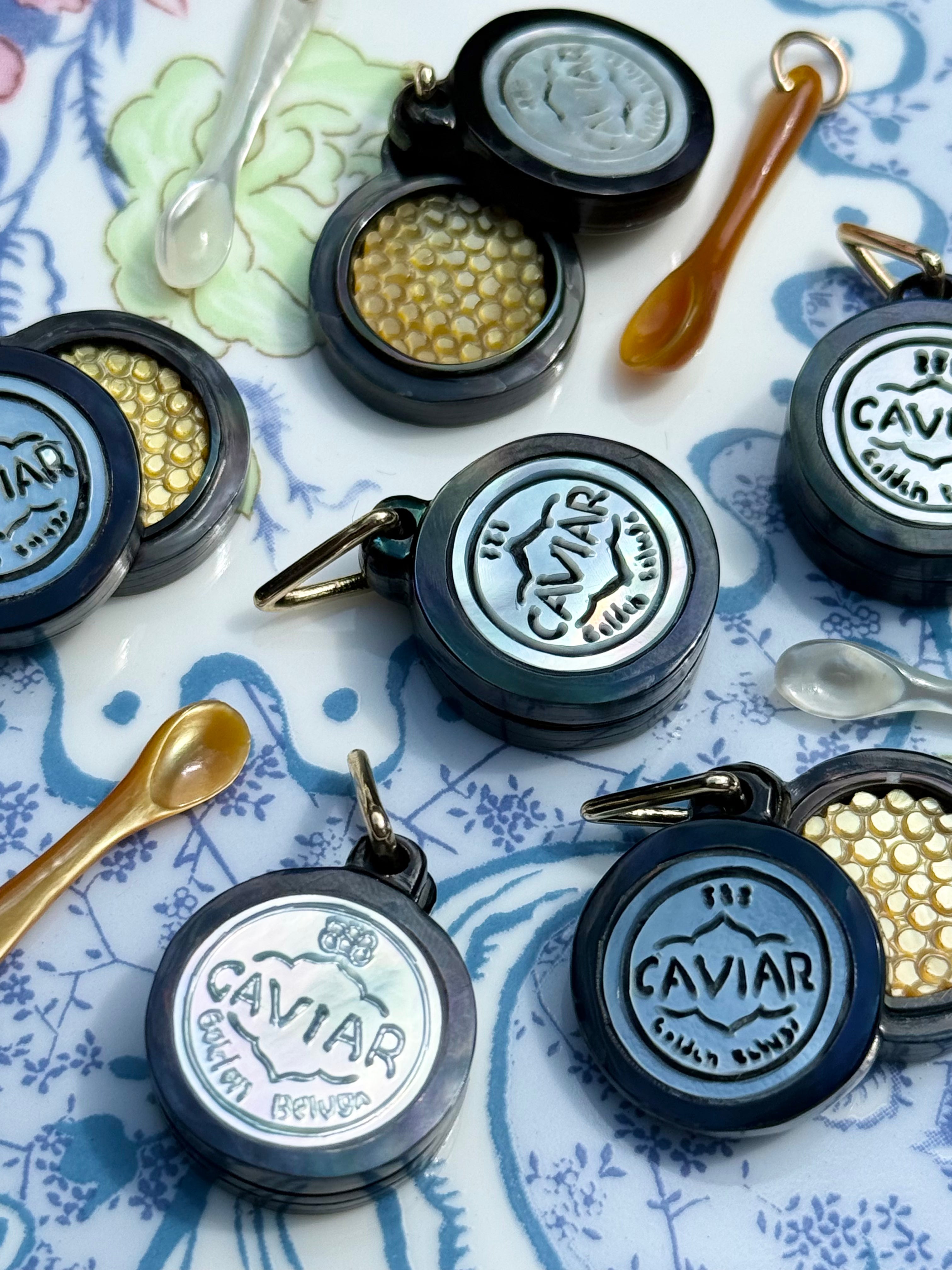 Several round black and silver caviar charms a floral surface, with small spoons placed around them. The lids are open to show gold caviar beads inside. Designed by Maura Green Jewelry