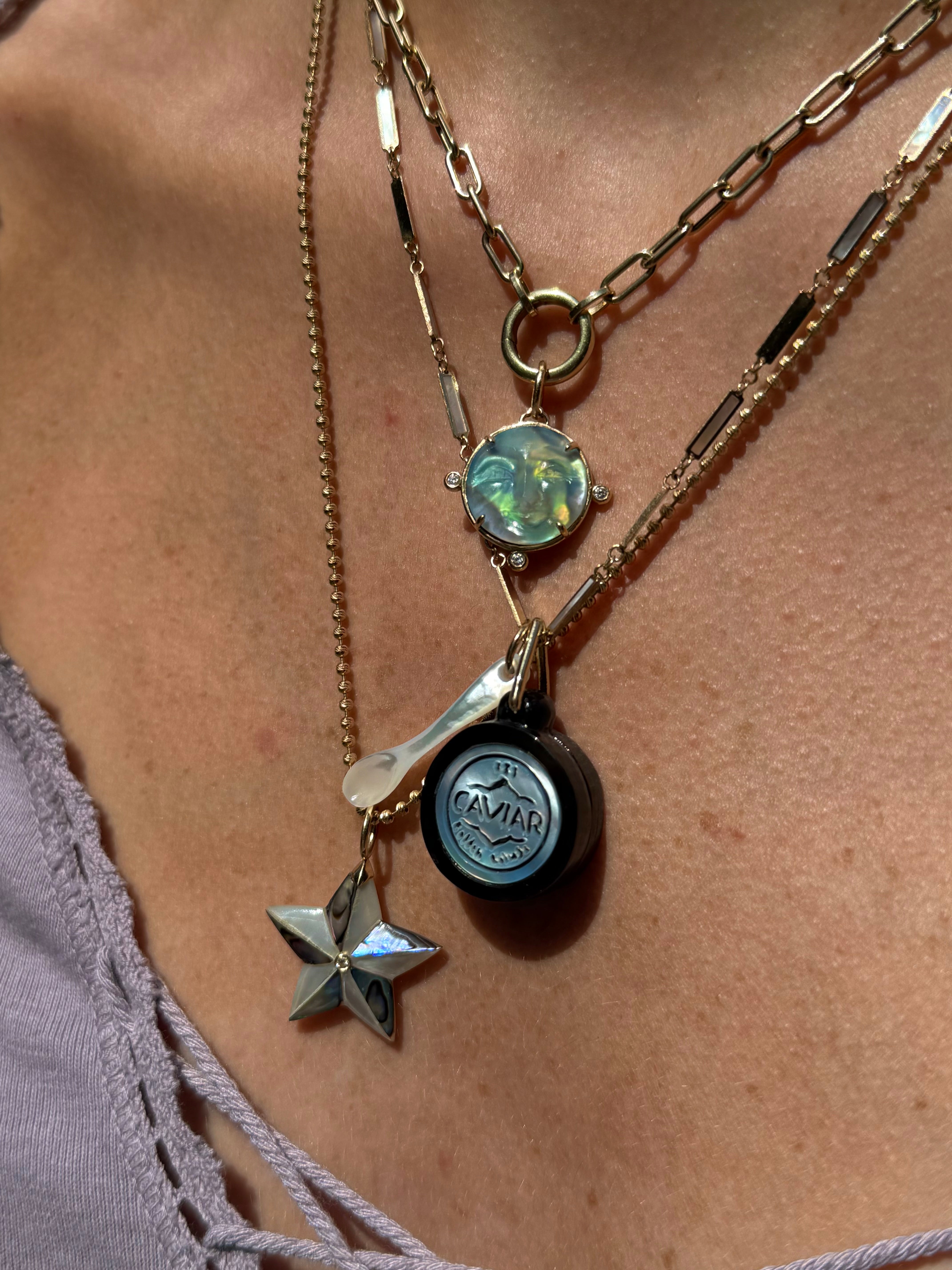 A close-up of a person's neck and chest wearing a layered gold-tone necklace set. The set includes a chain with a round iridescent moon face charm, a chain with a black round charm featuring a silver caviar design, and a separate luminescent star-shaped charm.