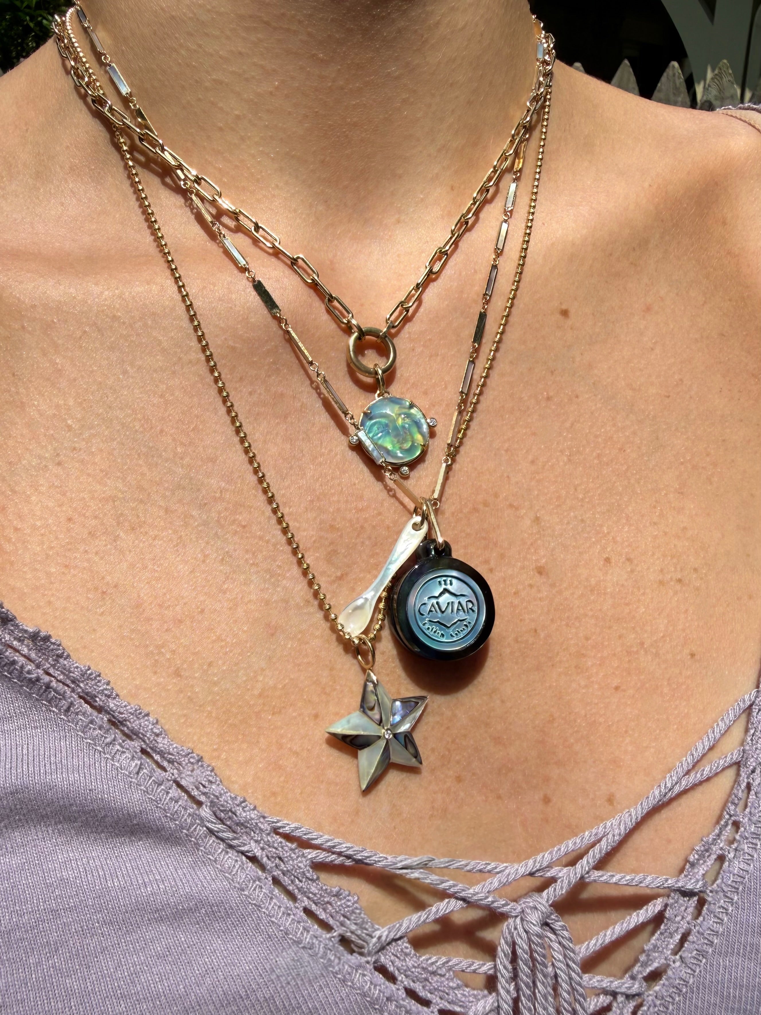 A close-up of a person's neck and chest wearing a layered necklace set. The necklace features multiple gold chains, including a star-shaped charm, a circular caviar charm, a luminescent moon face charm, and a star charm. Designed by Maura Green Jewelry.