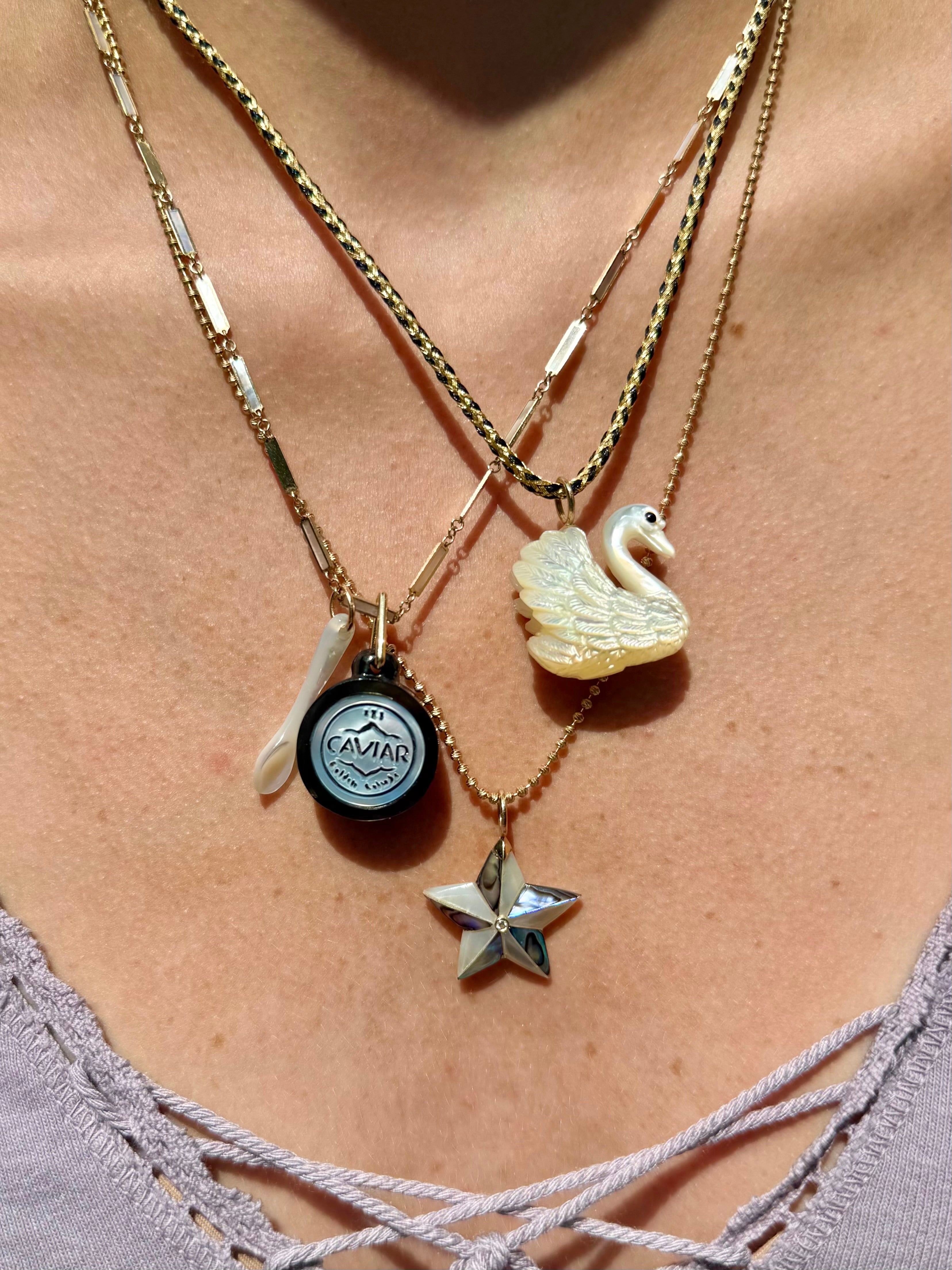 A close-up of a person wearing a layered necklace set with gold-toned chains, including a white swan pendant, a star pendant, a round black and white Caviar charm, and a small white spoon charm.