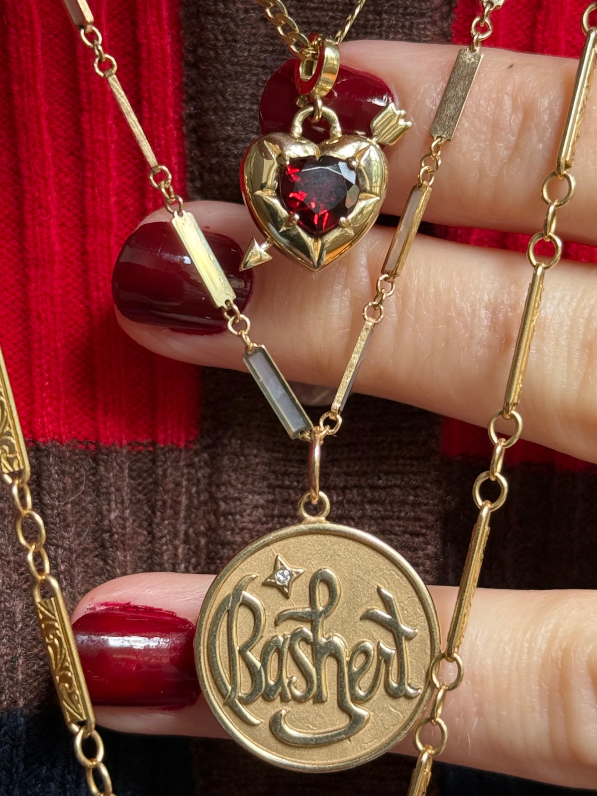 A close-up of a hand with dark red nail polish holding layered gold necklaces. One necklace features a Cupid's heart-shaped pendant with a red gemstone and arrow motif, while the lower pendant is a round gold medallion engraved with the word 'Bashert'. Designed by Maura Green