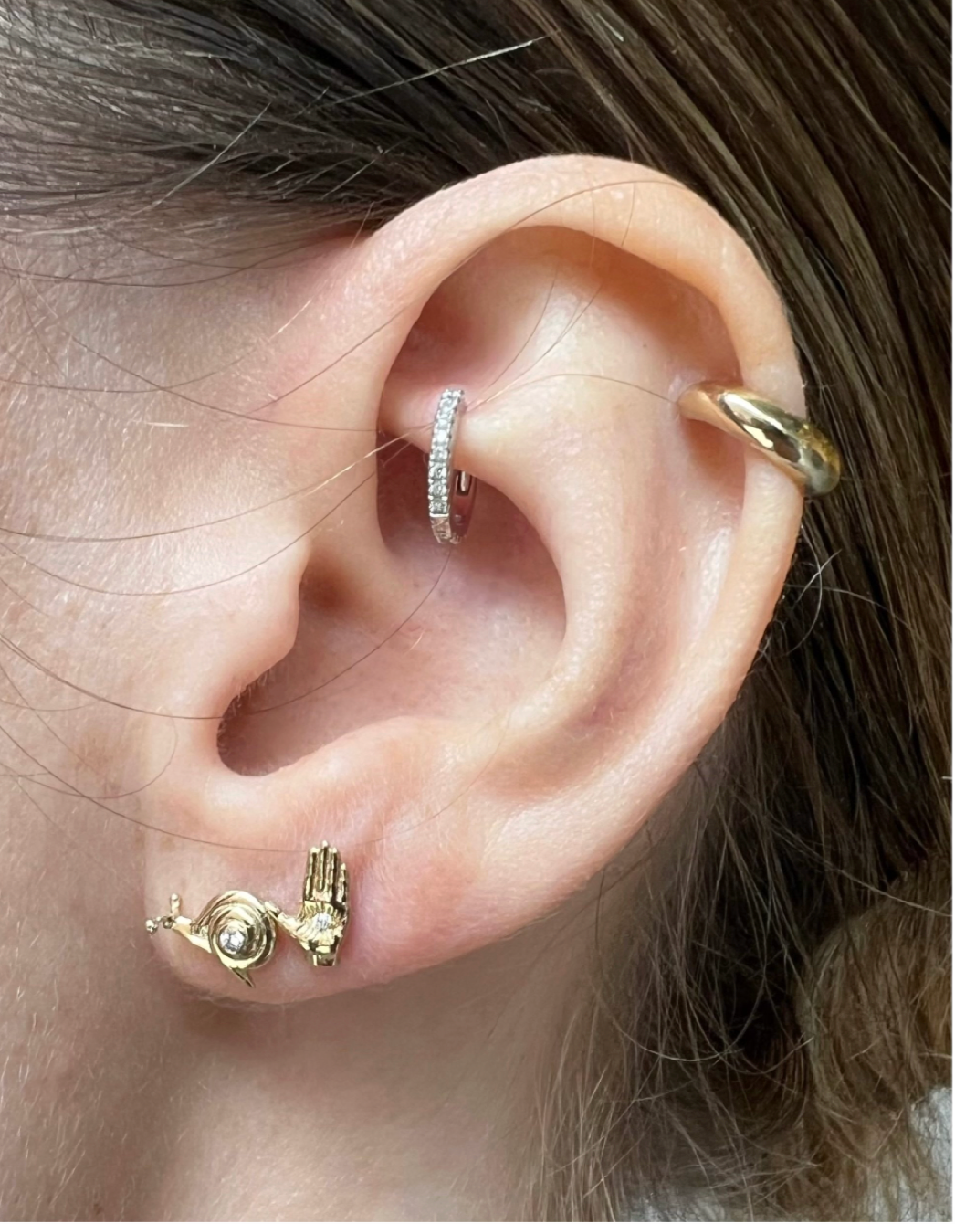 Close up of an ear with multiple piercings including Maura Green Jewelry gold snail stud, gold hamsa ladyfinger stud, gold huggie, and diamond huggie.