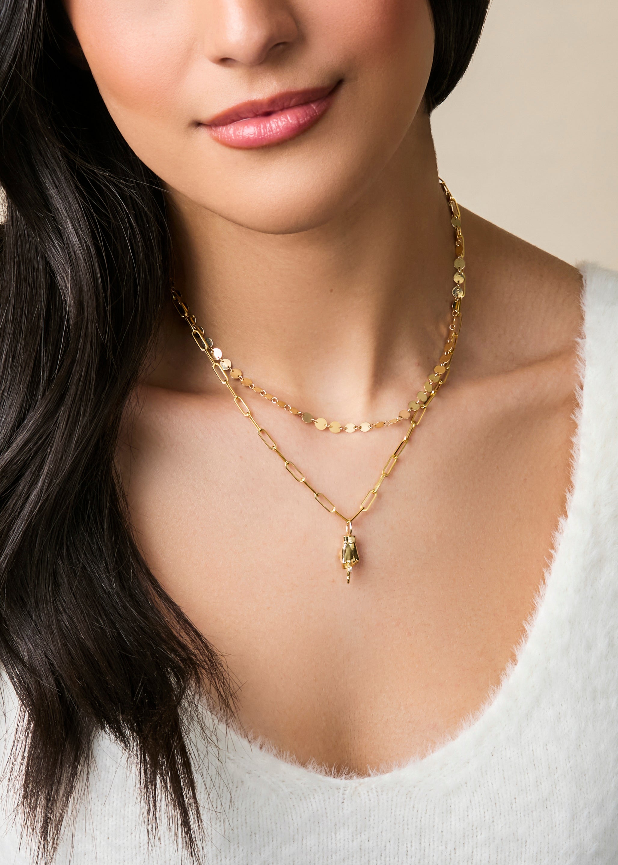 Cropped shot of a woman with long dark hair wearing a white shirt and two delicate gold layered necklaces. The bottom necklace has a gold figural hand charm in a middle finger gesture designed by Maura Green for her ladyfinger collection.