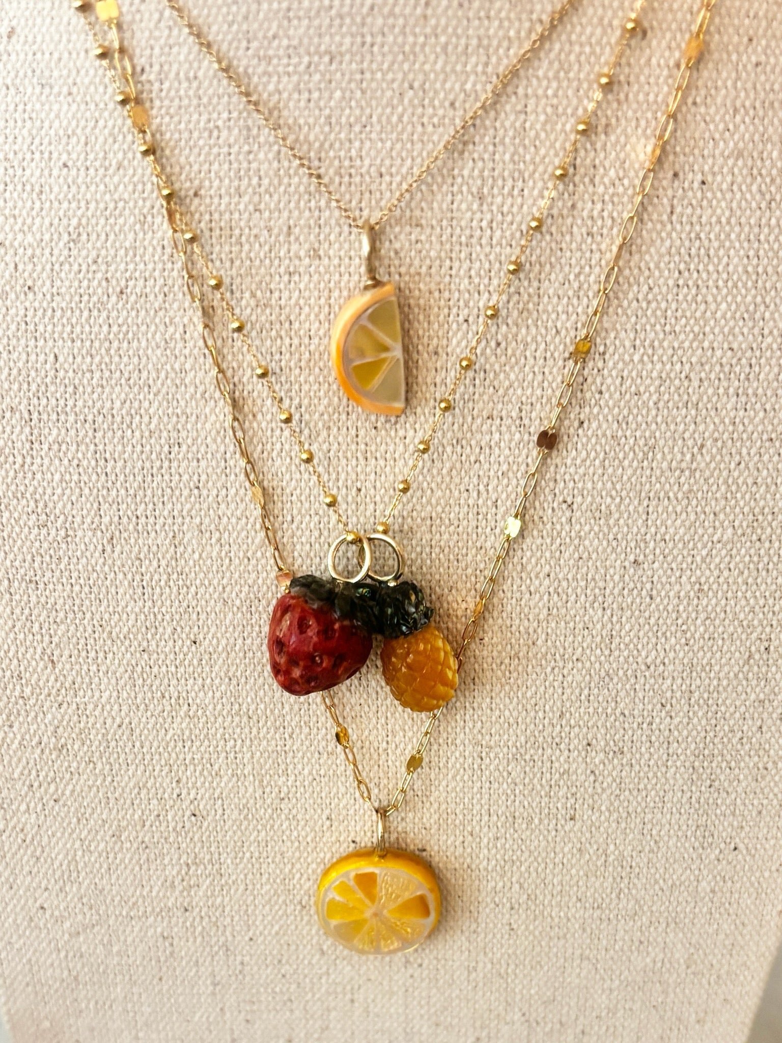 Layered fruit charm necklaces including an orange slice, a strawberry, a pineapple and a lemon slice against a beige linen background.