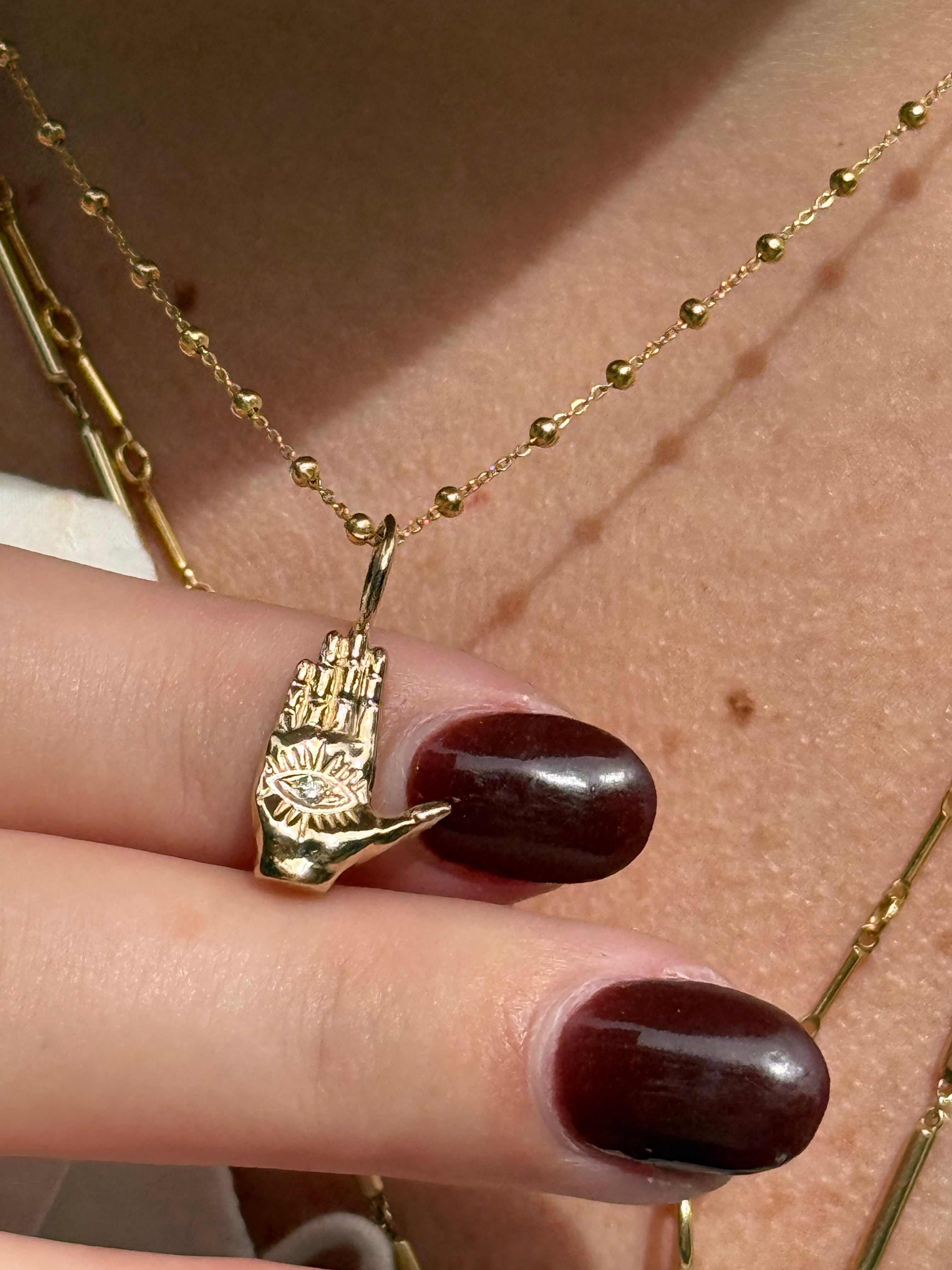 Gold figural hand hamsa charm on a finger with maroon nail polish, gold necklace in the background