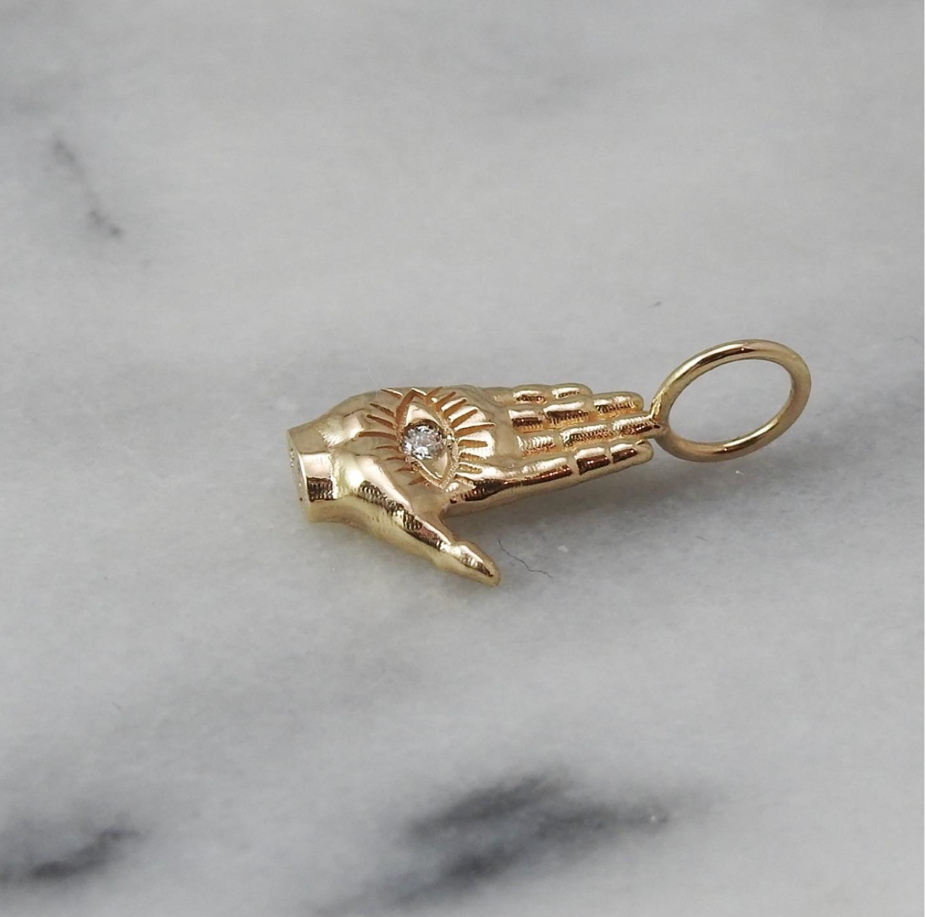 Maura Green Jewelry gold hamsa ladyfinger charm with a diamond in the palm of the hand on a white marble surface.