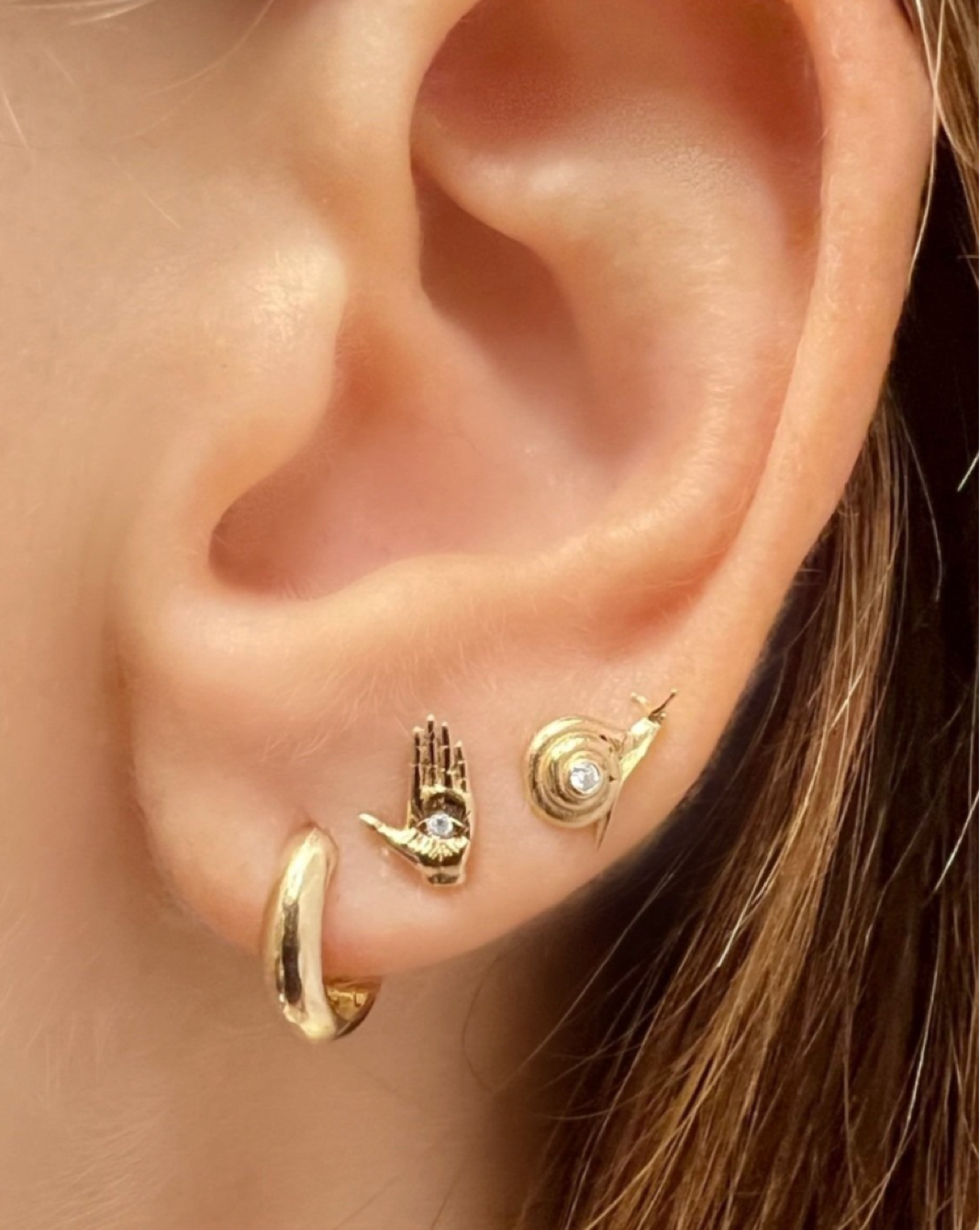 A close up of an ear stack including a gold huggie, and maura green hamsa ladyfinger stud in gold with a diamond in the center, and a gold snail stud with a diamond in the center