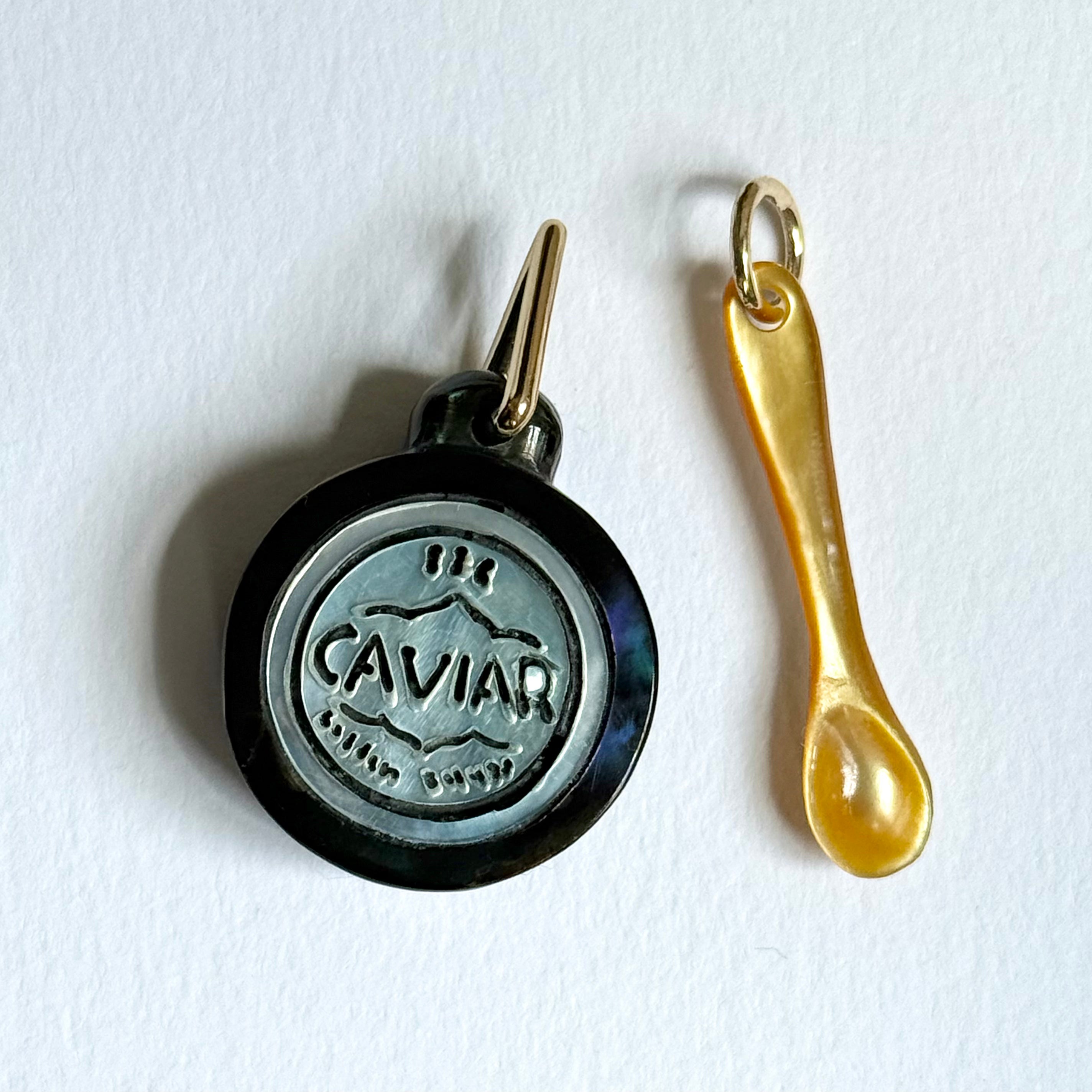 A gold mother of pearl caviar spoon charm and a round black and silver-toned caviar tin charm, displayed on a clean white background.  Designed by Maura Green Jewelry.