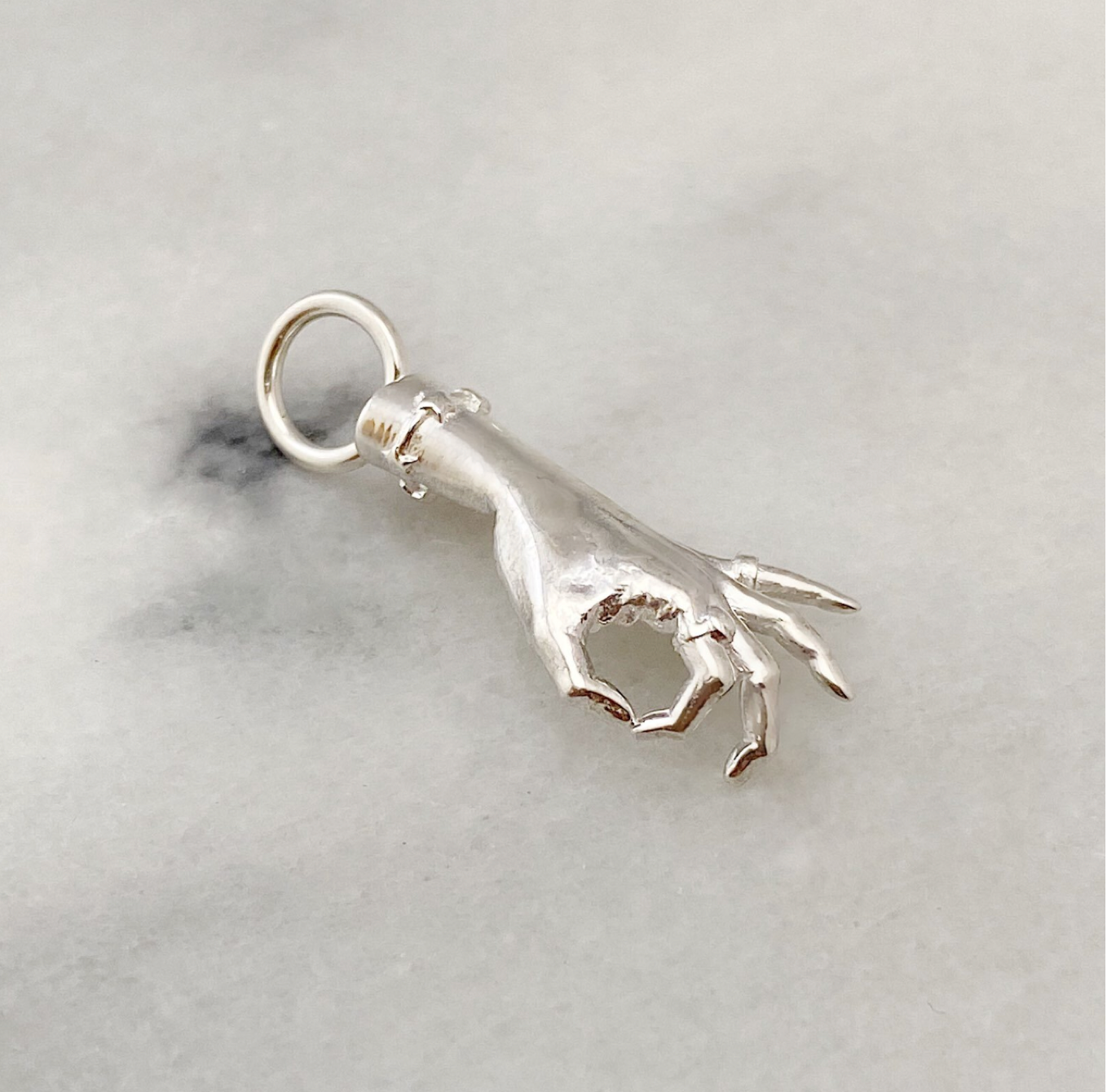 A silver colored sterling silver figural hand charm in the shape of the gyan mudra gesture with thumb and forefinger making a circle shape and the remaining 3 fingers raised. The charm is laying on a white marble surface and was designed by Maura Green Jewelry for her ladyfinger collection.
