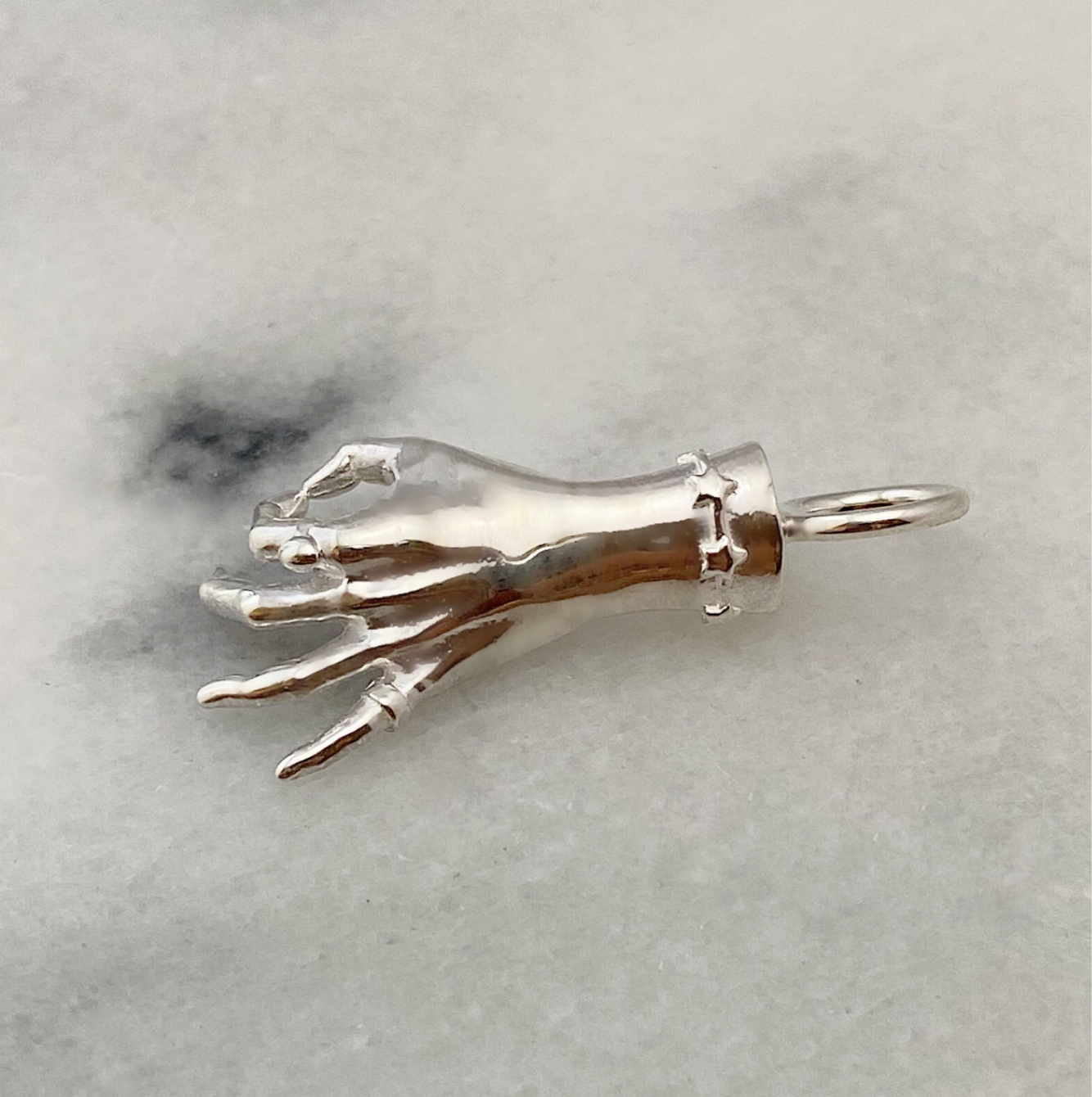 A silver colored sterling silver figural hand charm in the shape of the gyan mudra gesture with thumb and forefinger making a circle shape and the remaining 3 fingers raised. The charm is laying on a white marble surface and was designed by Maura Green Jewelry for her ladyfinger collection.