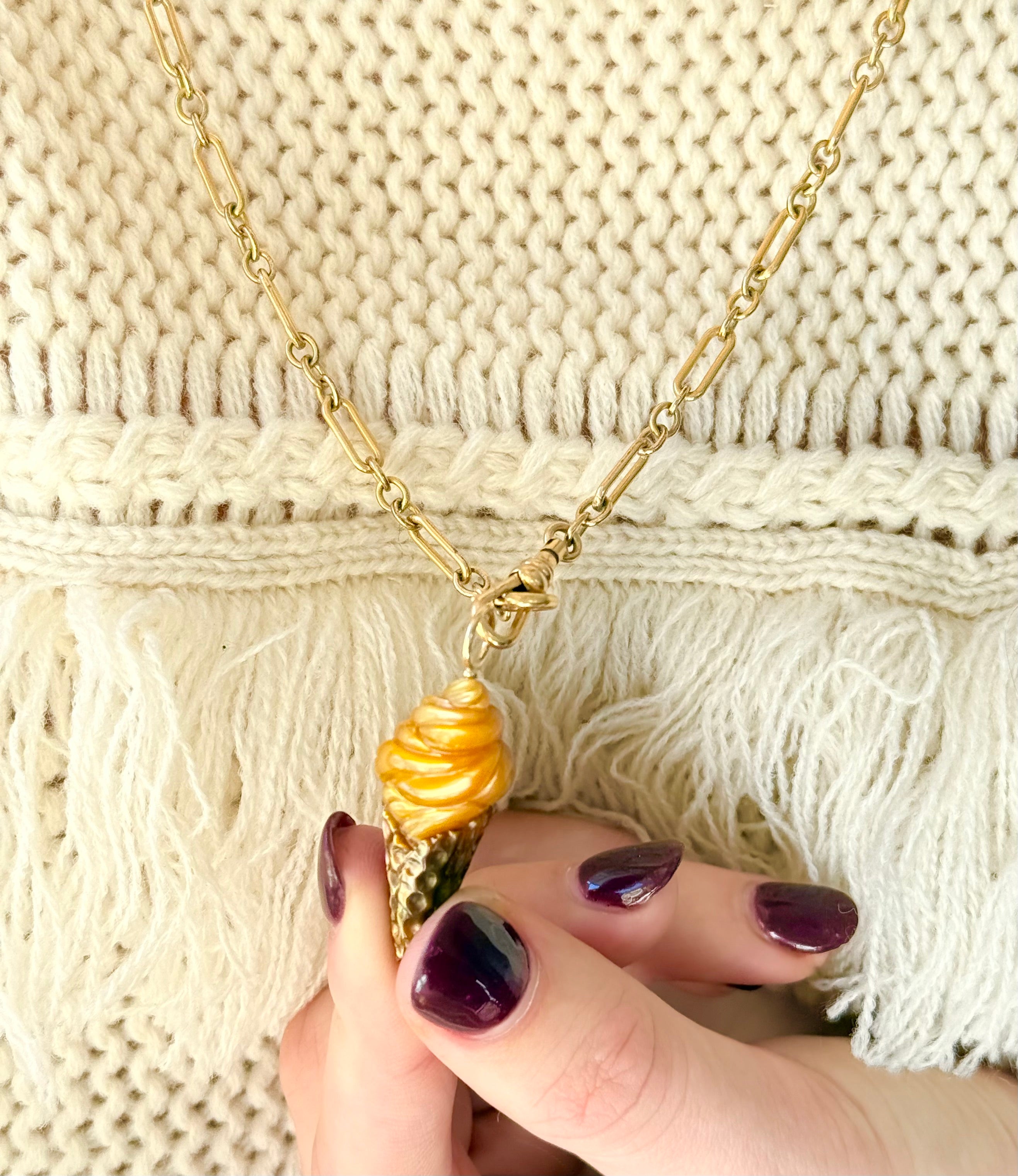 Gold ice cream conenecklace with a yellow pendant held by a hand against a textured beige background