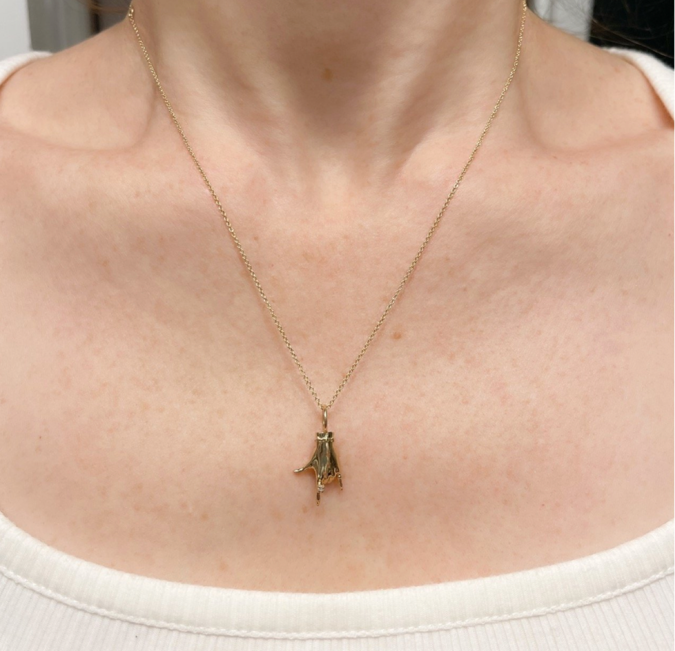 Cropped shot of a person wearing a delicate gold chain with a gold charm shaped like a hand making the American Sign Language symbol for I Love You designed by Maura Green Jewelry.