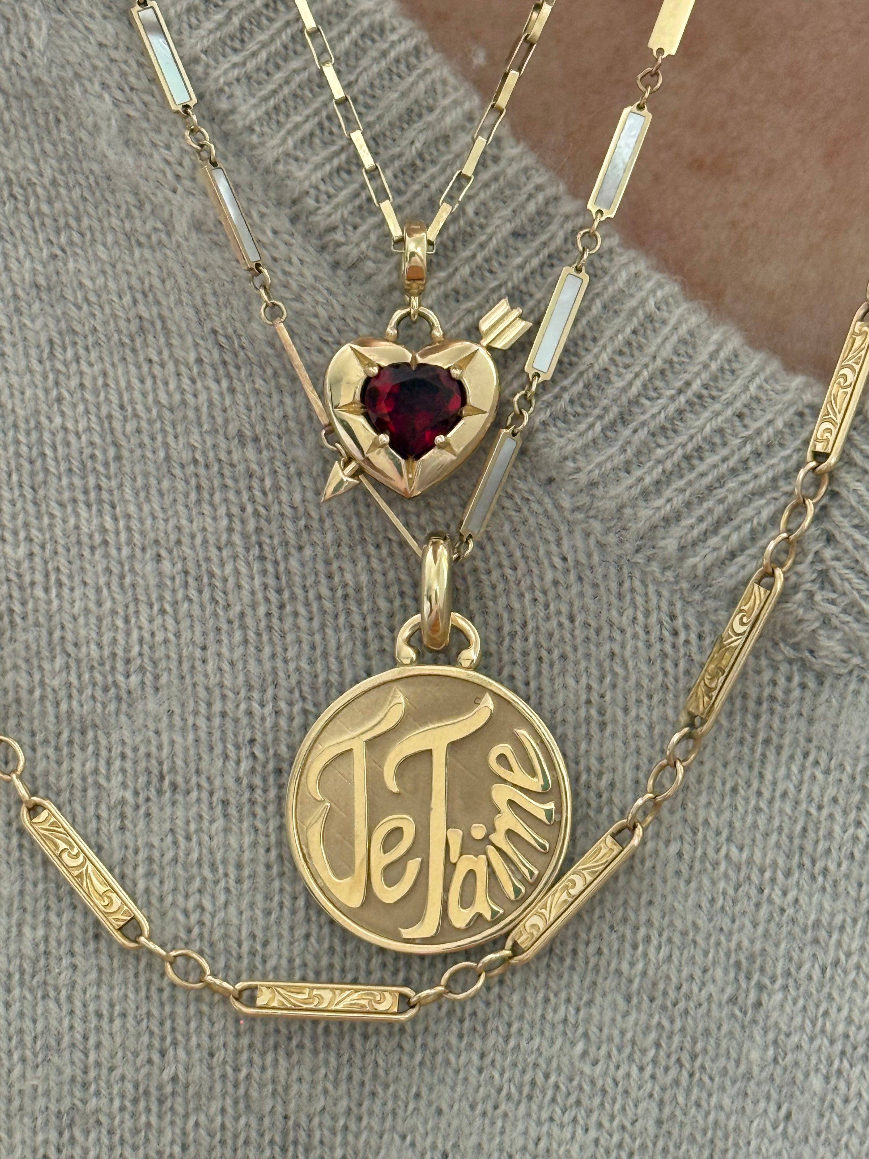 A gold-toned necklace with a round charm with "Je T'aime" engraved, a heart-shaped charm with a dark red gemstone, Designed by Maura Green Jewelry