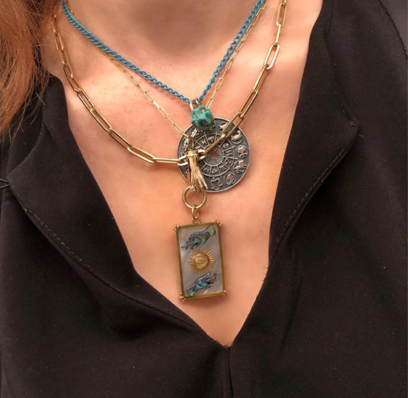 Close up shot of several layered necklaces designed by Maura Green on a woman wearing a black shirt. The necklaces include a blackened silver zodiac wheel charm on a blue chain, and rectangular blue, gold  and white charm representing the sun tarot card on a gold chain.