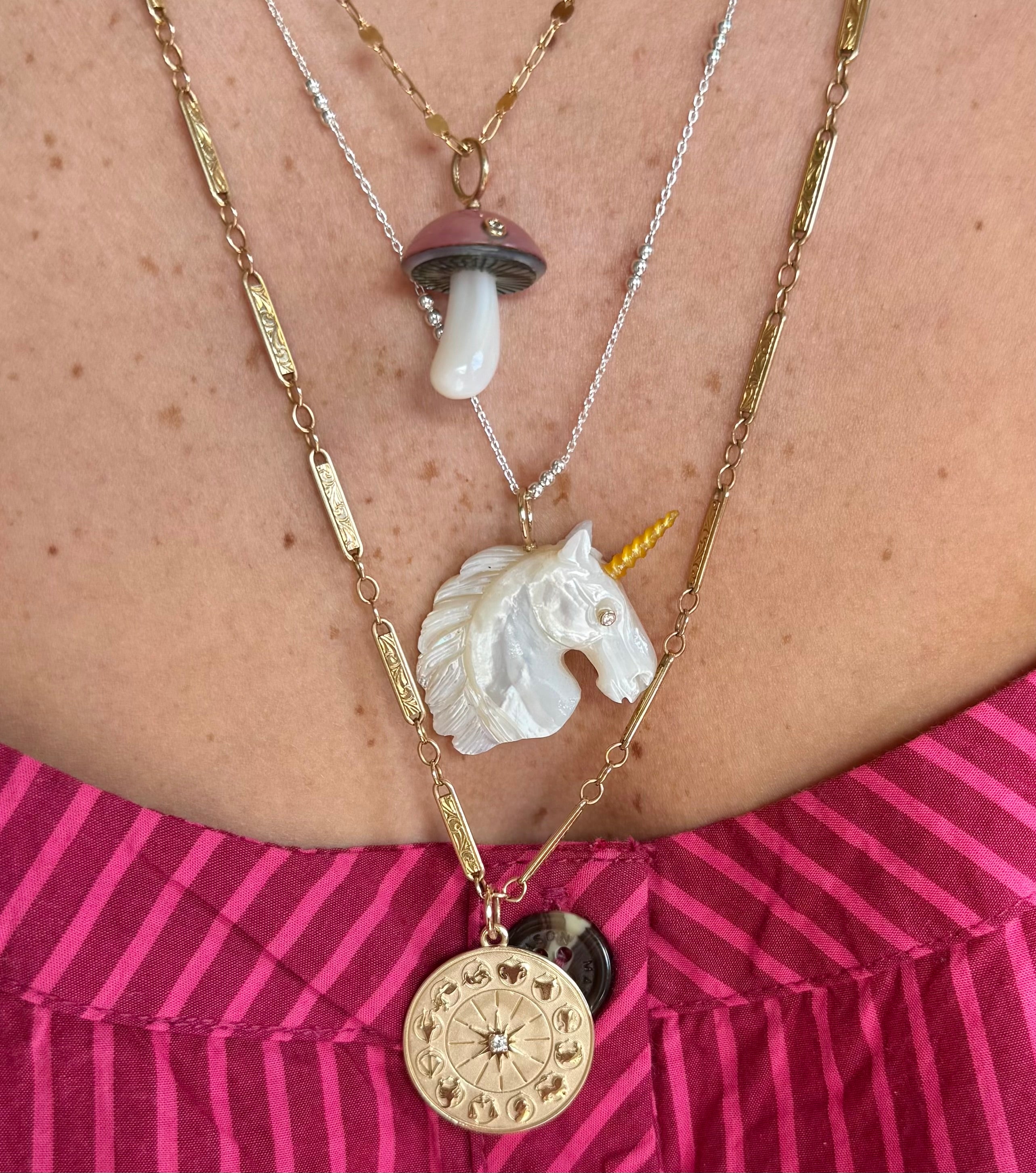 Close-up of a person wearing multiple necklaces with unique pendants including one of a pink mushroom, one of a white unicorn, and one of a zodiac wheel in gold.