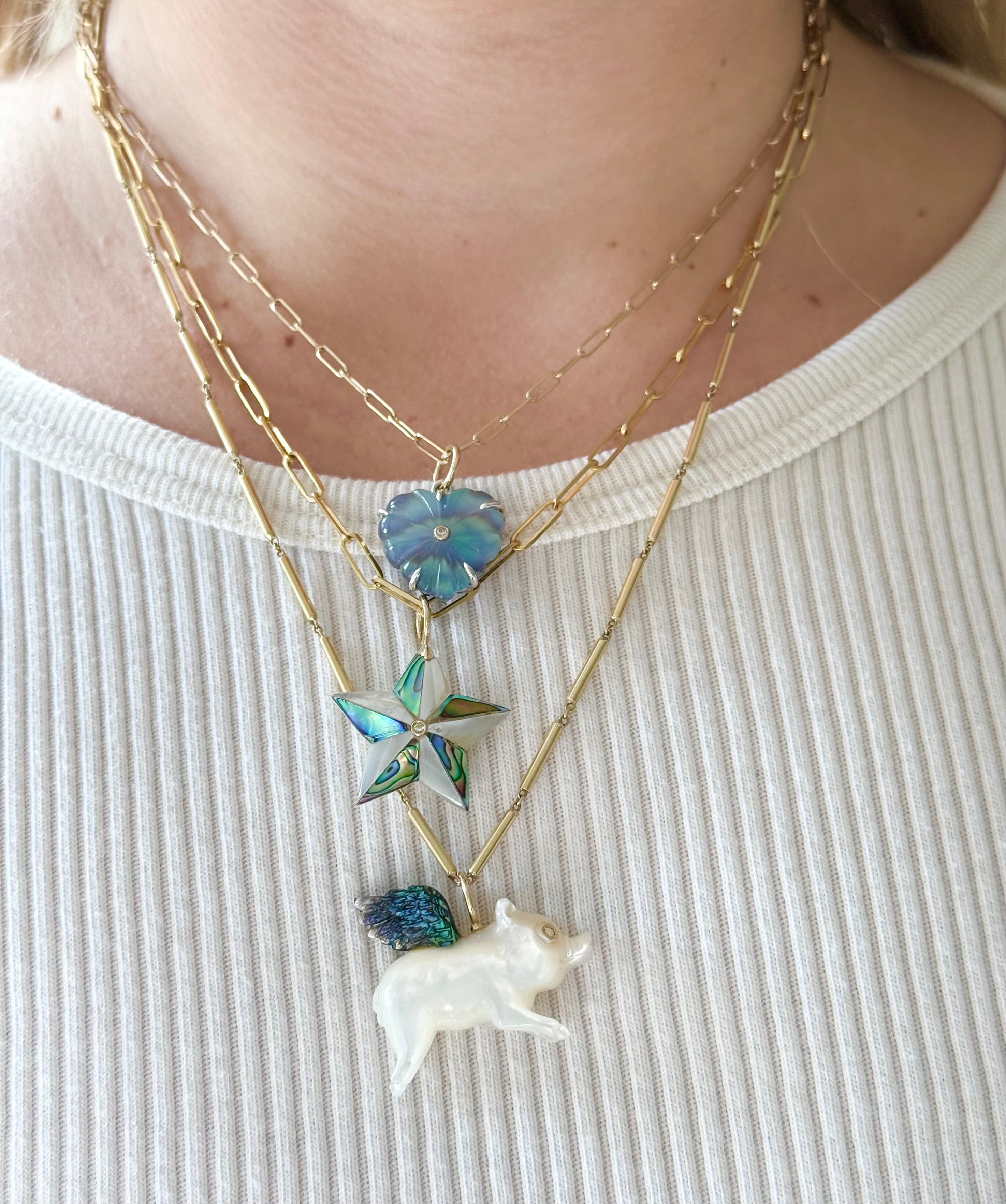 Necklaces with a white flying pig pendant and a blue star pendant, and a blue heart shaped pendant on a person wearing a white top.