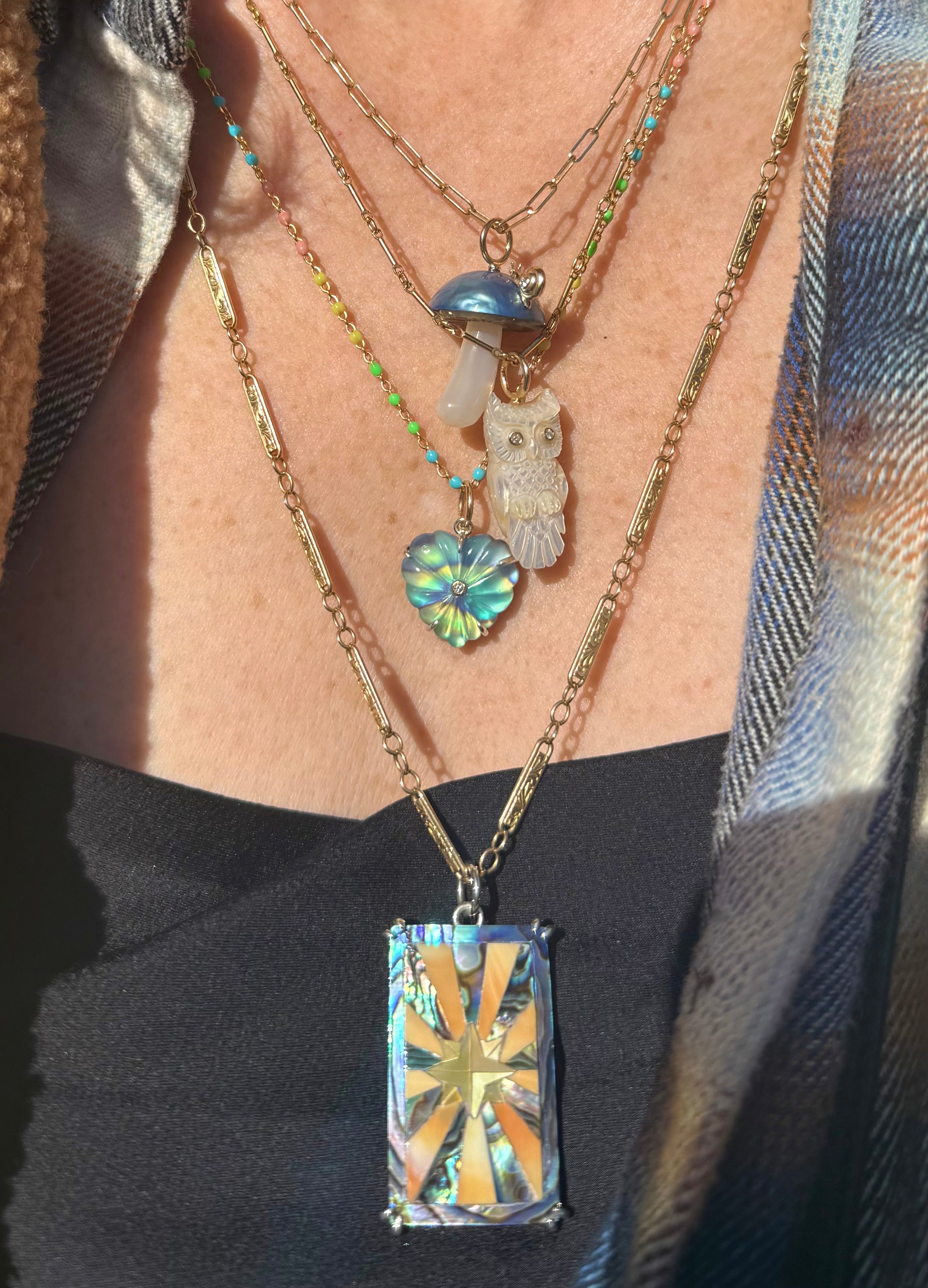 A layered series of necklaces with various pendants including a white owl charm, a blue mushroom charm, heart-shaped charm in blue and green, and a rectangular charm featuring a starburst.