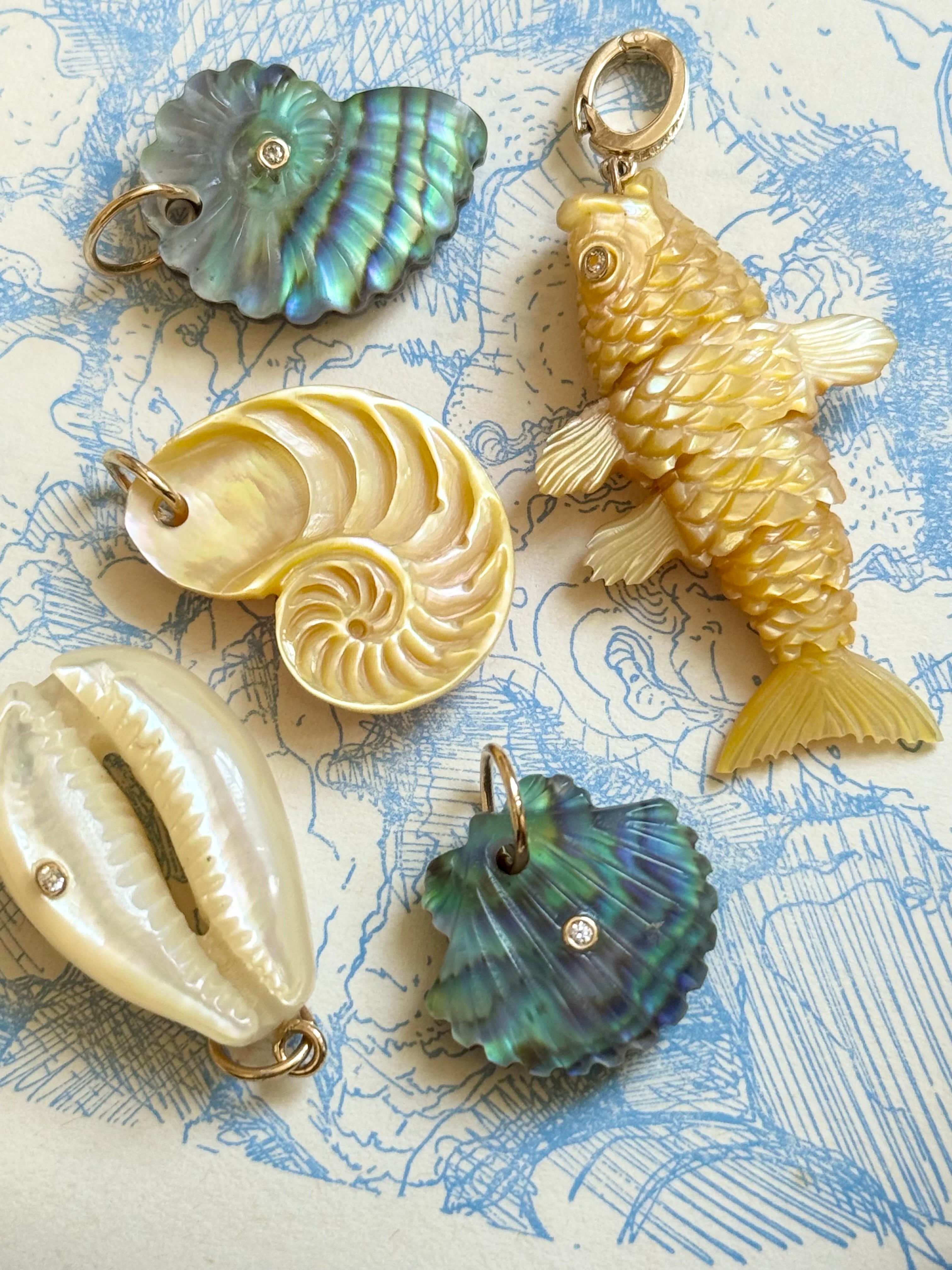 Collection of Maura Green Jewelry shell and fish-shaped pendants on a textured blue and white surface.