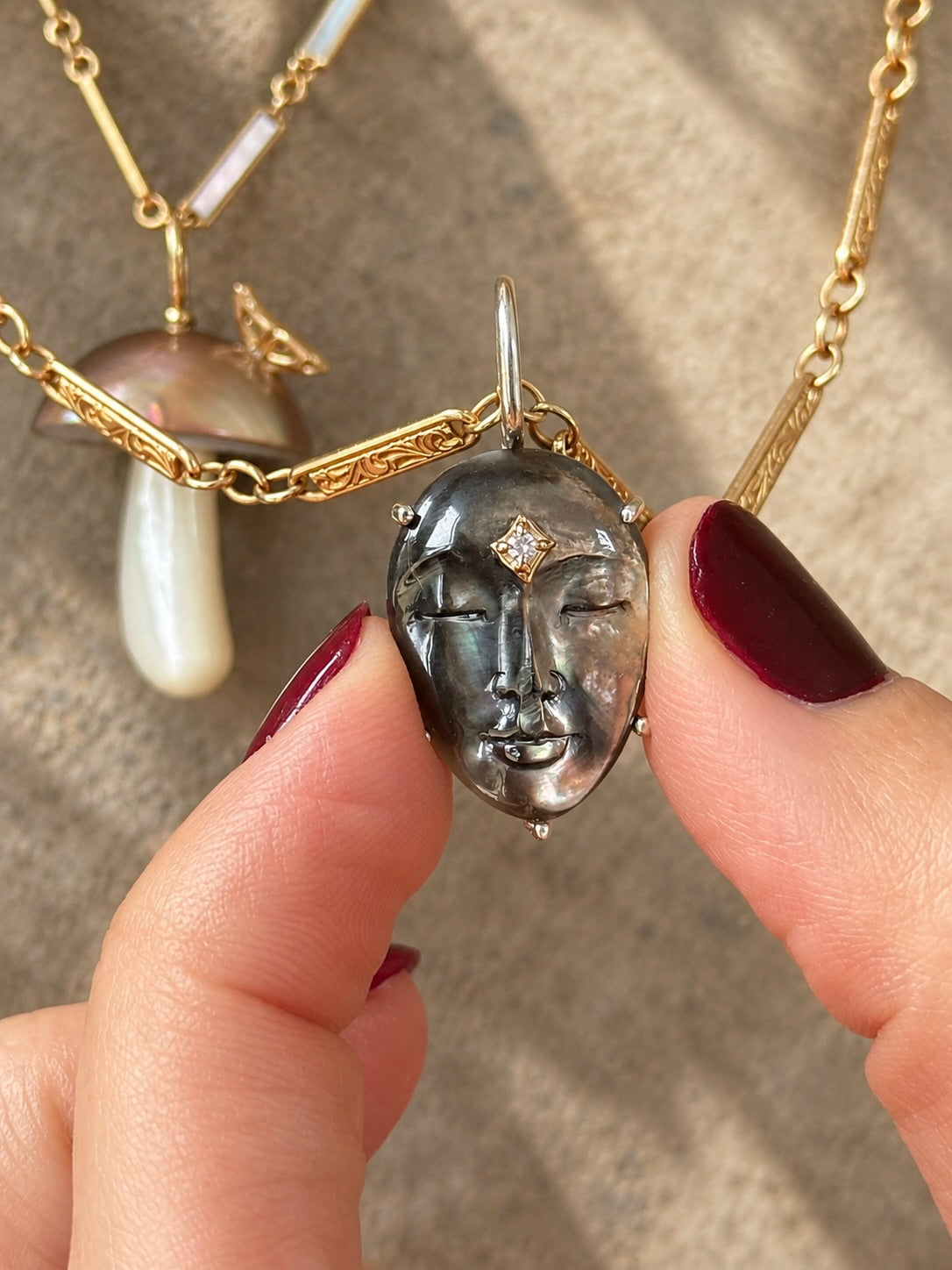 Close-up of a hand holding a carved black mother-of-pearl face charm accented with a diamond and set on a mixed 14K gold and mother-of-pearl chain, with a mushroom charm in the background.