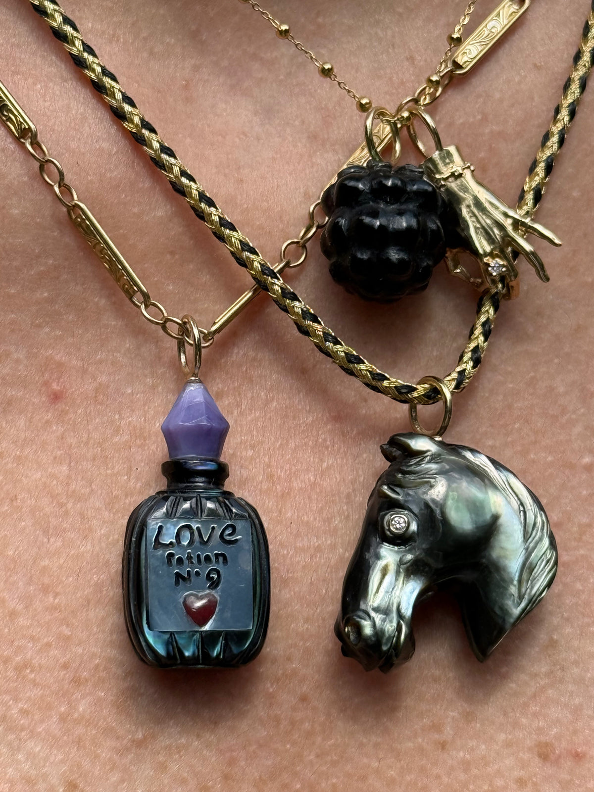 A close-up of a person's skin wearing layered necklaces. The necklaces include a gold and black braided chain with a black berry charm, a gold chain with a small gold charm, and a gold chain with a dark metallic horse head pendant and a dark bottle pendant with a purple cap.