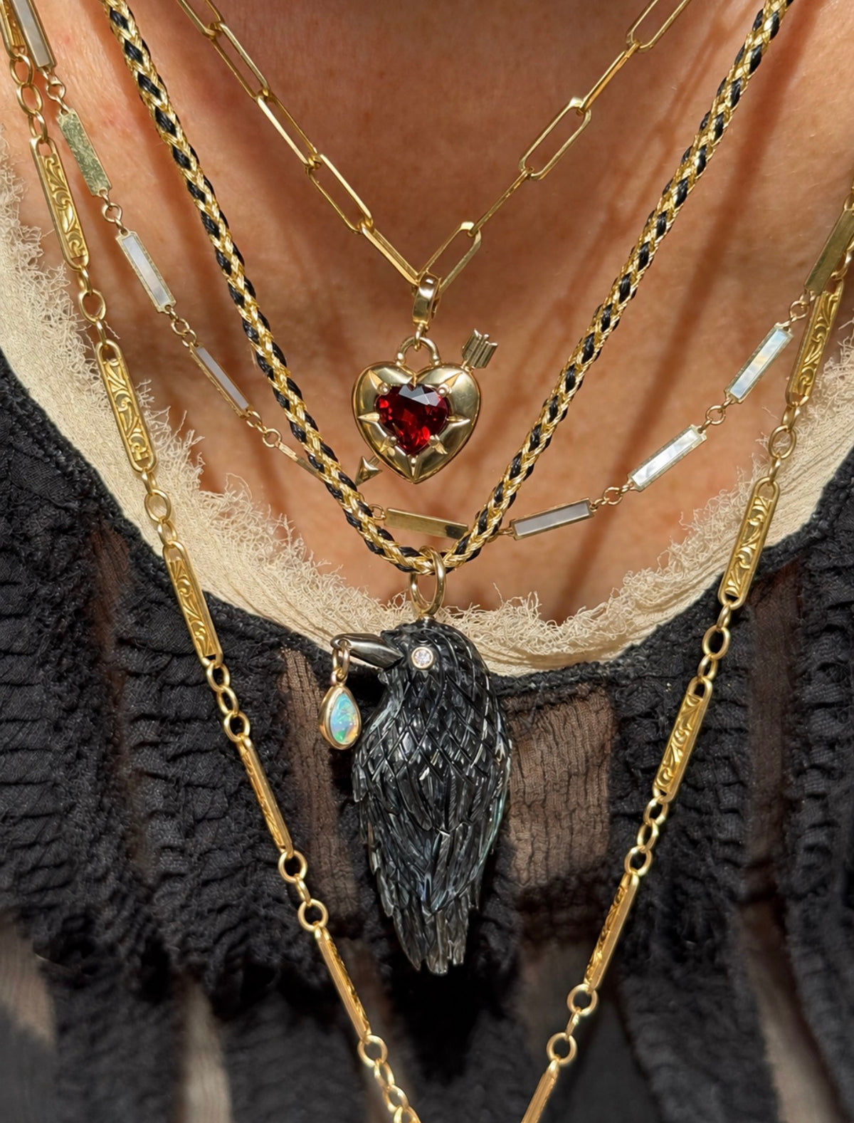 A close-up of a person wearing layered gold necklaces, including a heart-shaped pendant with a red gemstone and a raven or crow pendant with a small blue teardrop charm, over a black and beige top.