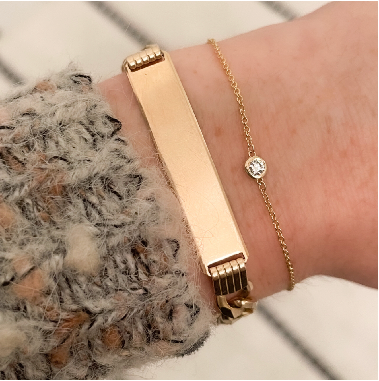 A woman's wrist showing the cuff of a sweater showing off two gold bracelets including a gold ID bracelet with a high polish metal finish, and a delicate gold chain bracelet with a bezel set diamond at it's center.