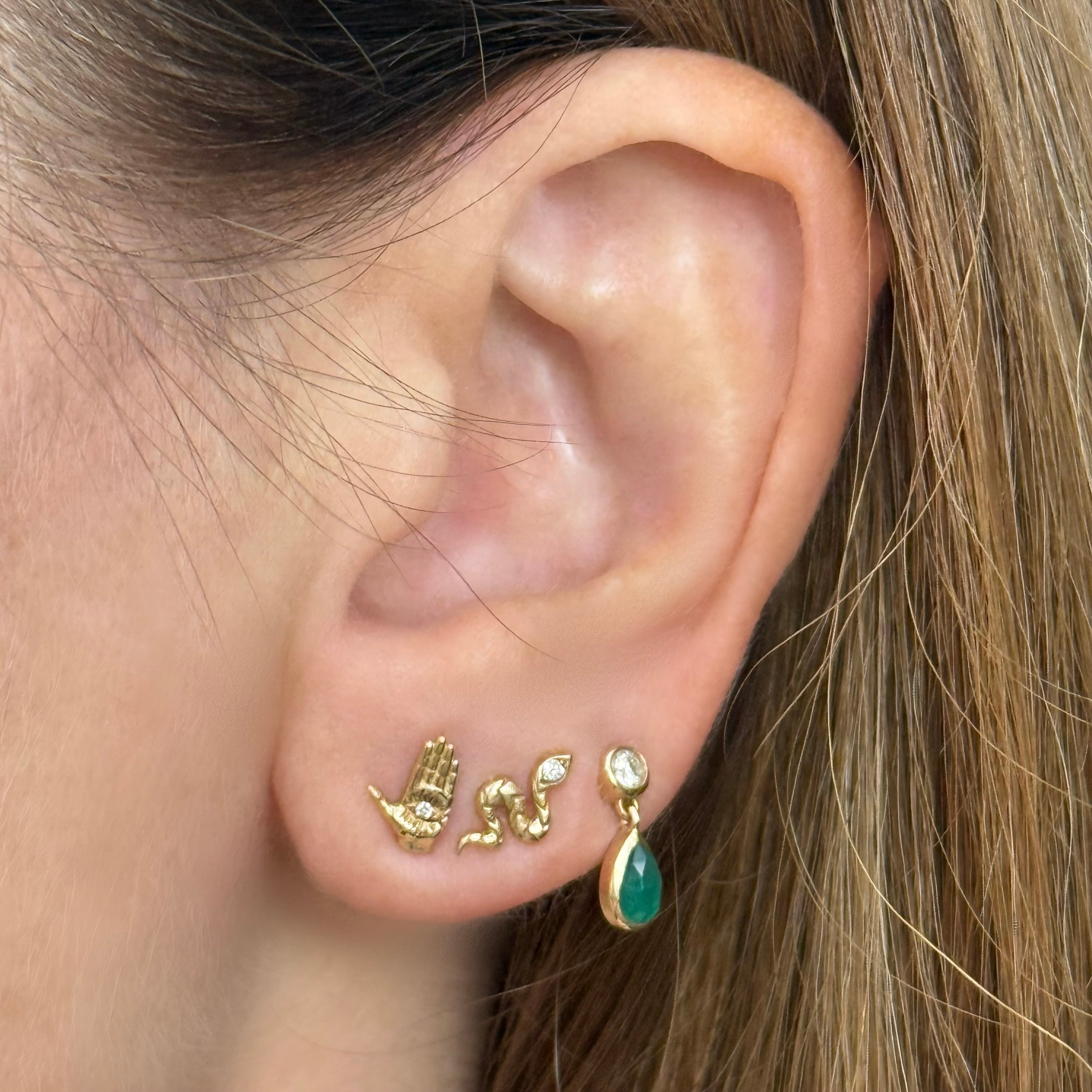 Close-up of an ear wearing Maura Green Jewelry gold stud earrings including the hamsa ladyfinger stud, the snake stud, and the emerald and diamond drop dangle stud. 