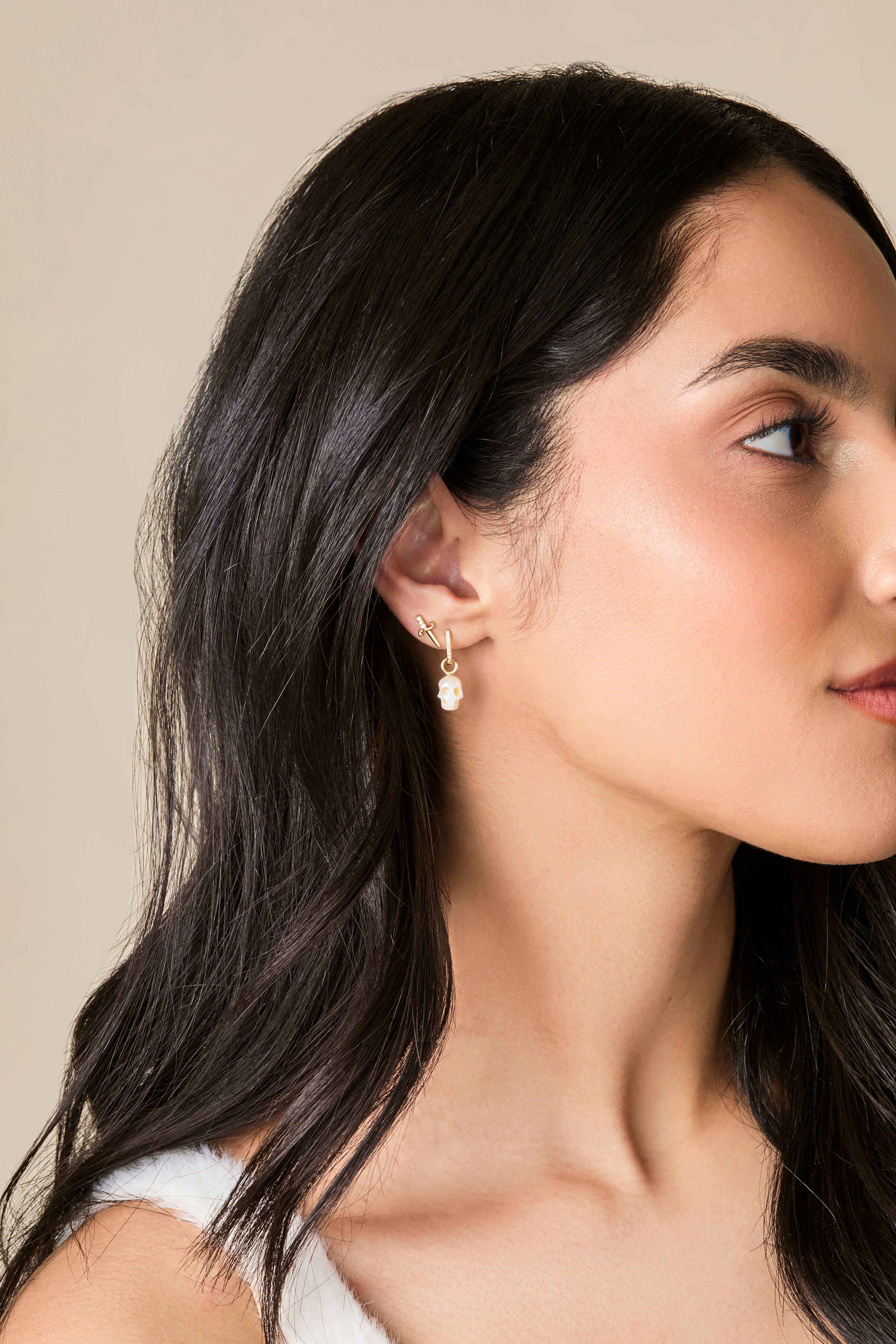 Profile of a model with long black hair wearing earrings by Maura Green Jewelry, including a diamond and gold dagger stud and a pearl skull dangling from a diamond and gold huggie.