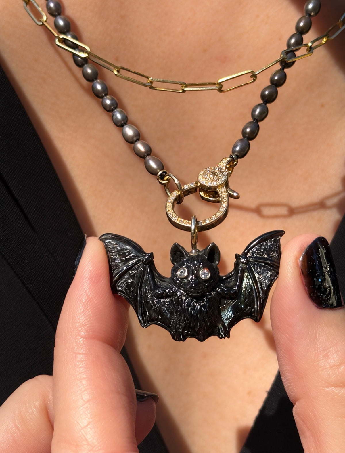 Close-up of a Maura Green Jewelry hand-carved black pinna shell bat charm with outstretched wings, diamond eyes, and a 14K yellow gold bail. The charm hangs from a diamond-accented gold clasp on a dark gray pearl necklace, layered with a gold paperclip chain. A model holds the bat between two fingers with black polished nails.