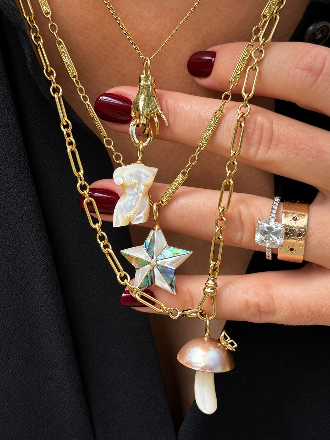 A close-up of a person's hand wearing a layered gold necklace set with unique charms, including a hand pendant, a shell star, a mushroom charm, and a paperclip pendant, paired with a black blazer.