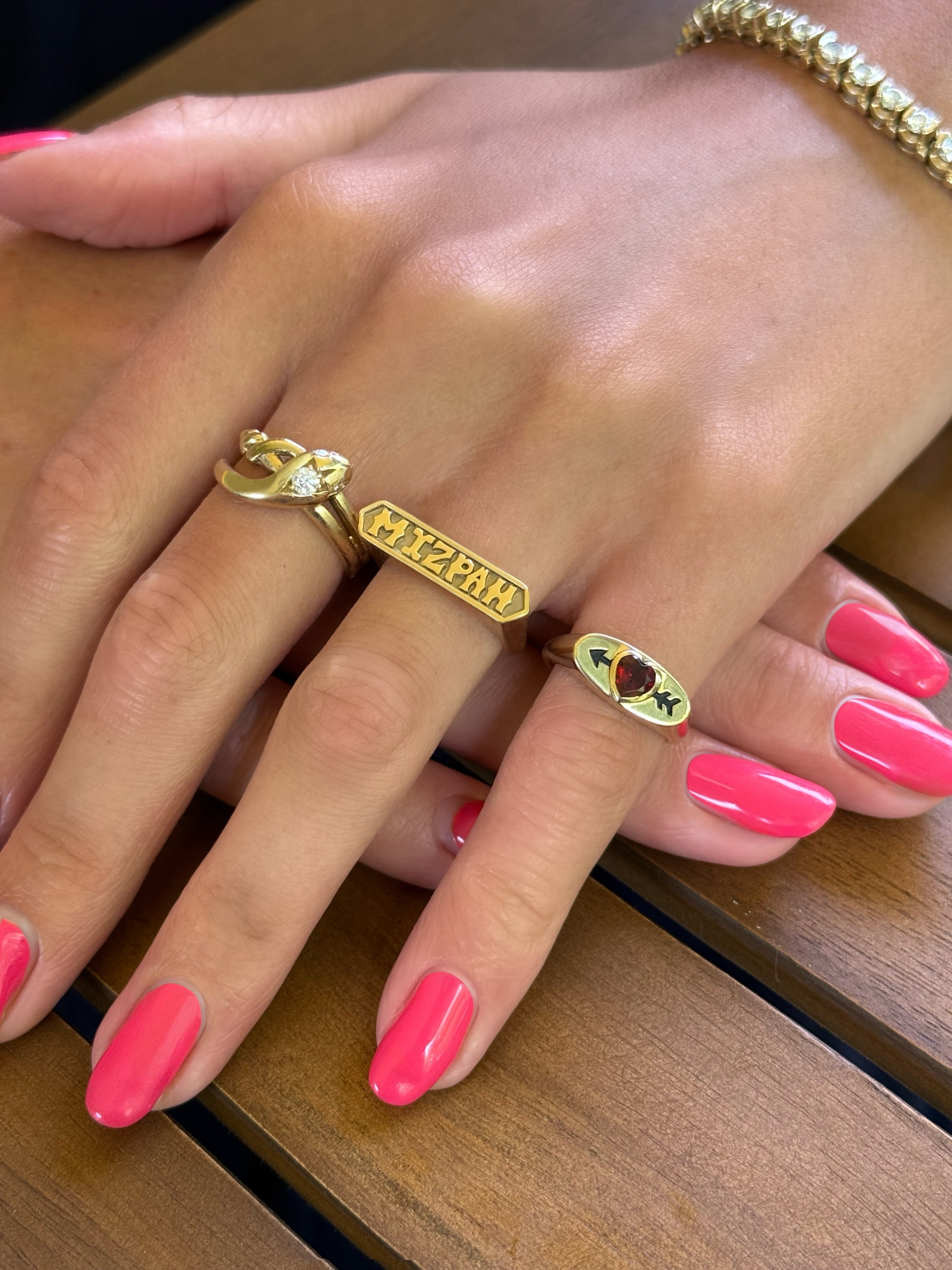 A woman's hands with a pink manicure wearing three gold rings designed by Maura Green, including a snake ring with a diamond on it's head, a mizpah ring, and a signet ring with a garnet heart and black enamel arrow shooting through it.