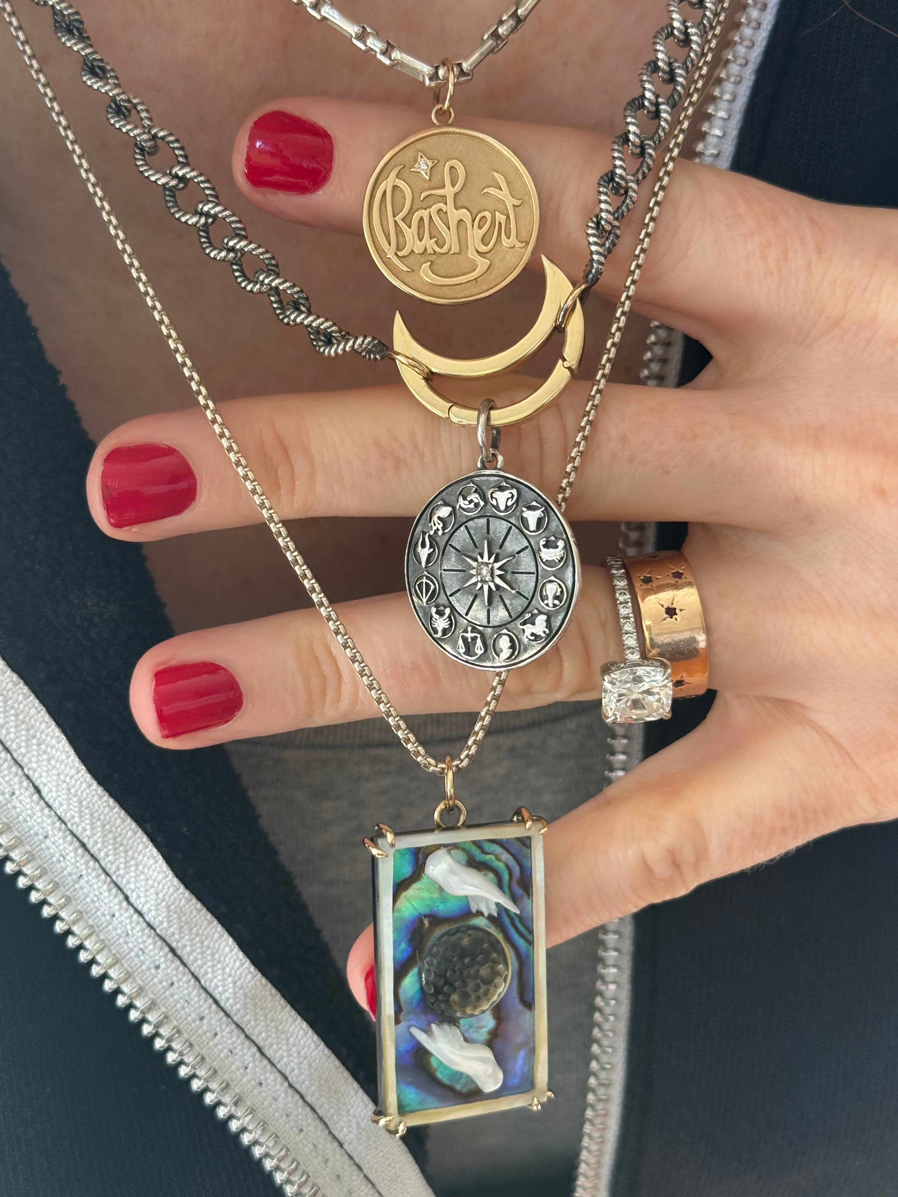 Hand wearing multiple necklaces with various pendants, including a 'Bashert' pendant designed by Maura Green Jewelry.