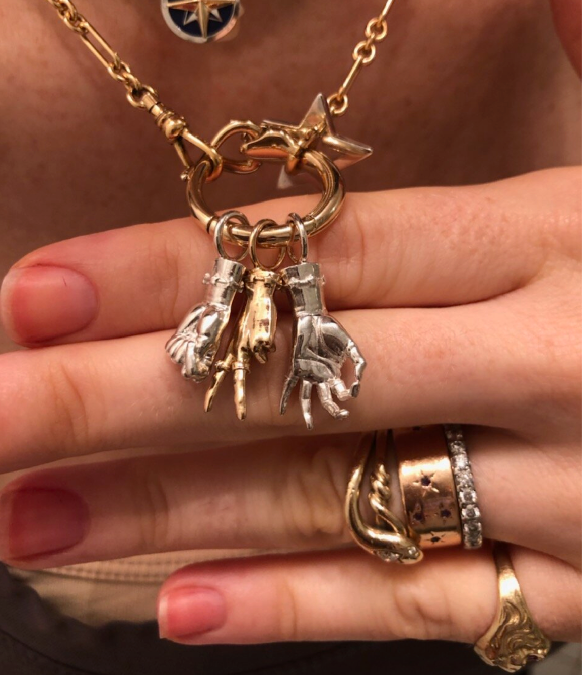 Close up of multiple Maura Green Jewelry ladyfinger charms on a circular charm holder leaning on a person's hand. The charms include a silver mudra shaped figural hand, a gold peace sign shaped figural hand, and a silver mano figa shaped figural hand.