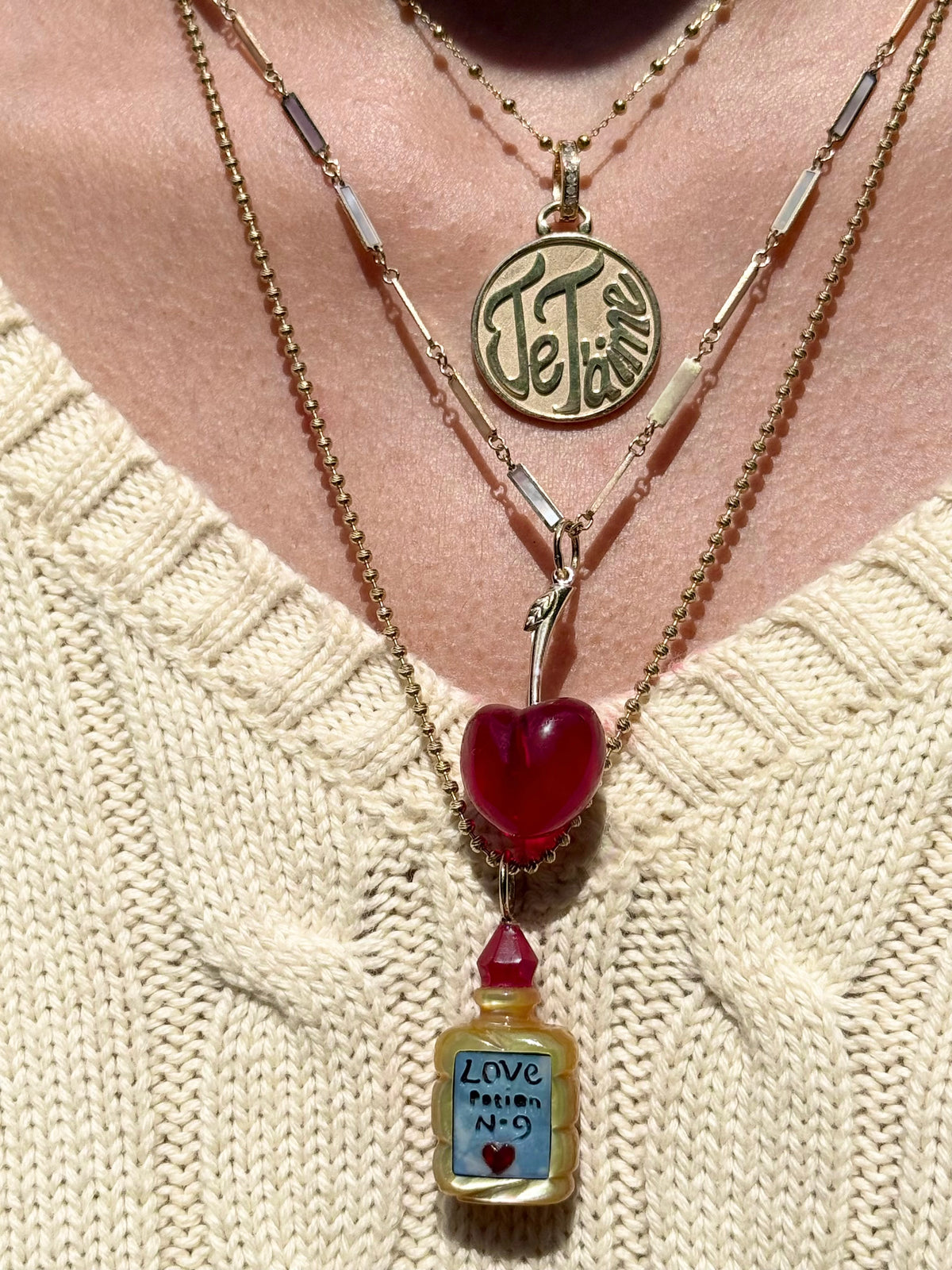 A close-up of a person wearing layered necklaces over a cream knit sweater. The necklaces include a round gold pendant which says "Je Taime", a red chalcedony cherry charm ona gold stem, and a small bottle pendant labeled 'Love Potion N-9'.