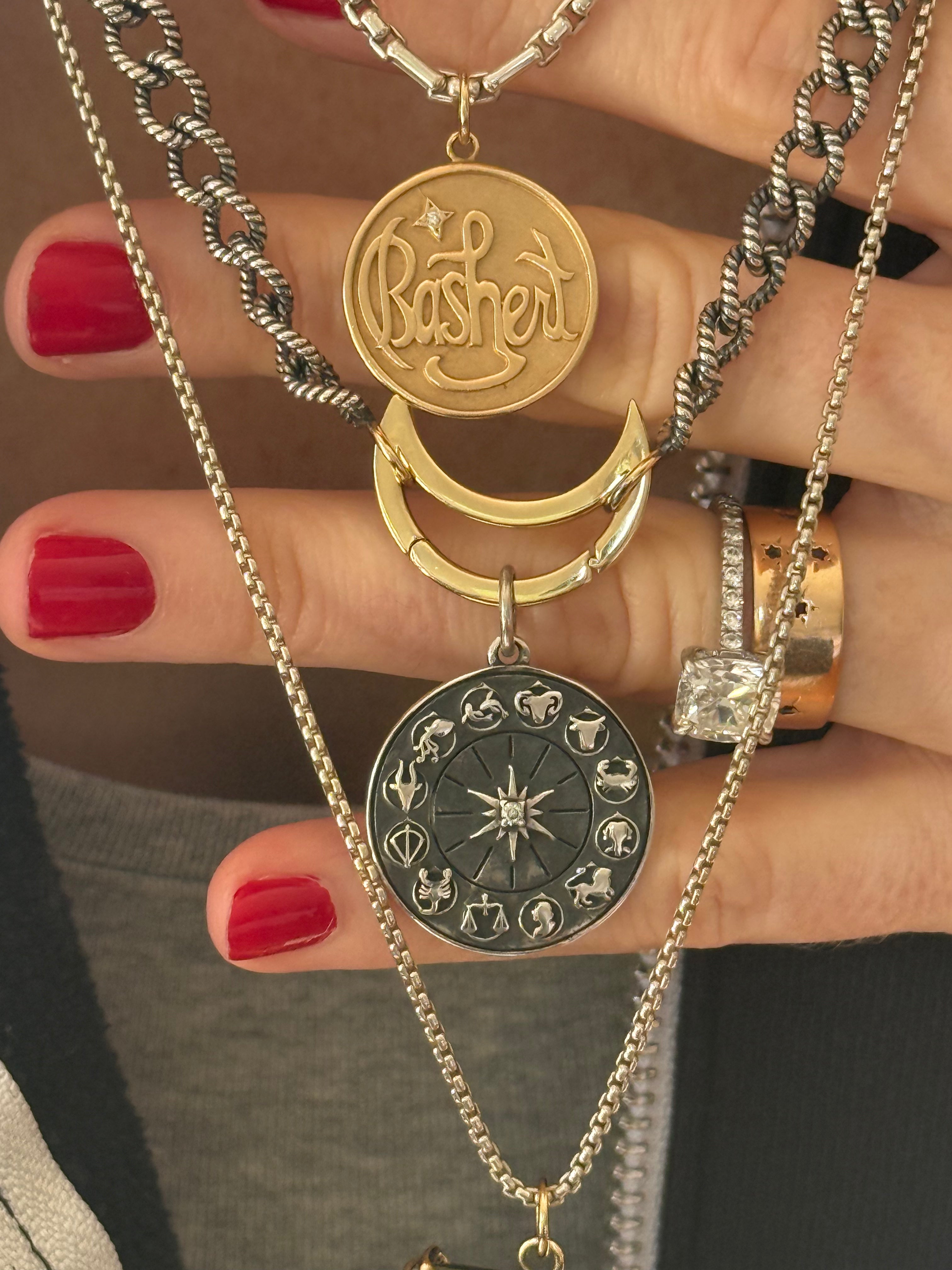 Gold and silver necklaces with pendants including a "bashert" charm and silver zodiac held by a hand with red nail polish designed by Maura Green Jewelry.