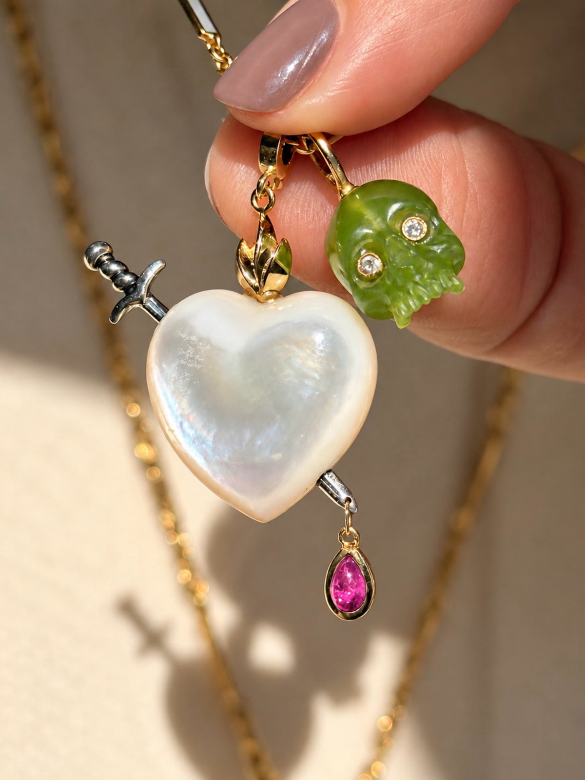 A taupe manicured forefinger and thumb gold up a white mother of pearl Sacred heart charm which is pierced by a silver sword, and has gold flames and a dangling teardrop shaped ruby at the bottom of the tip of the sword. Next to the sacred heart is a grass green chalcedony carved skull charm with diamond eyes. Designed by Maura Green