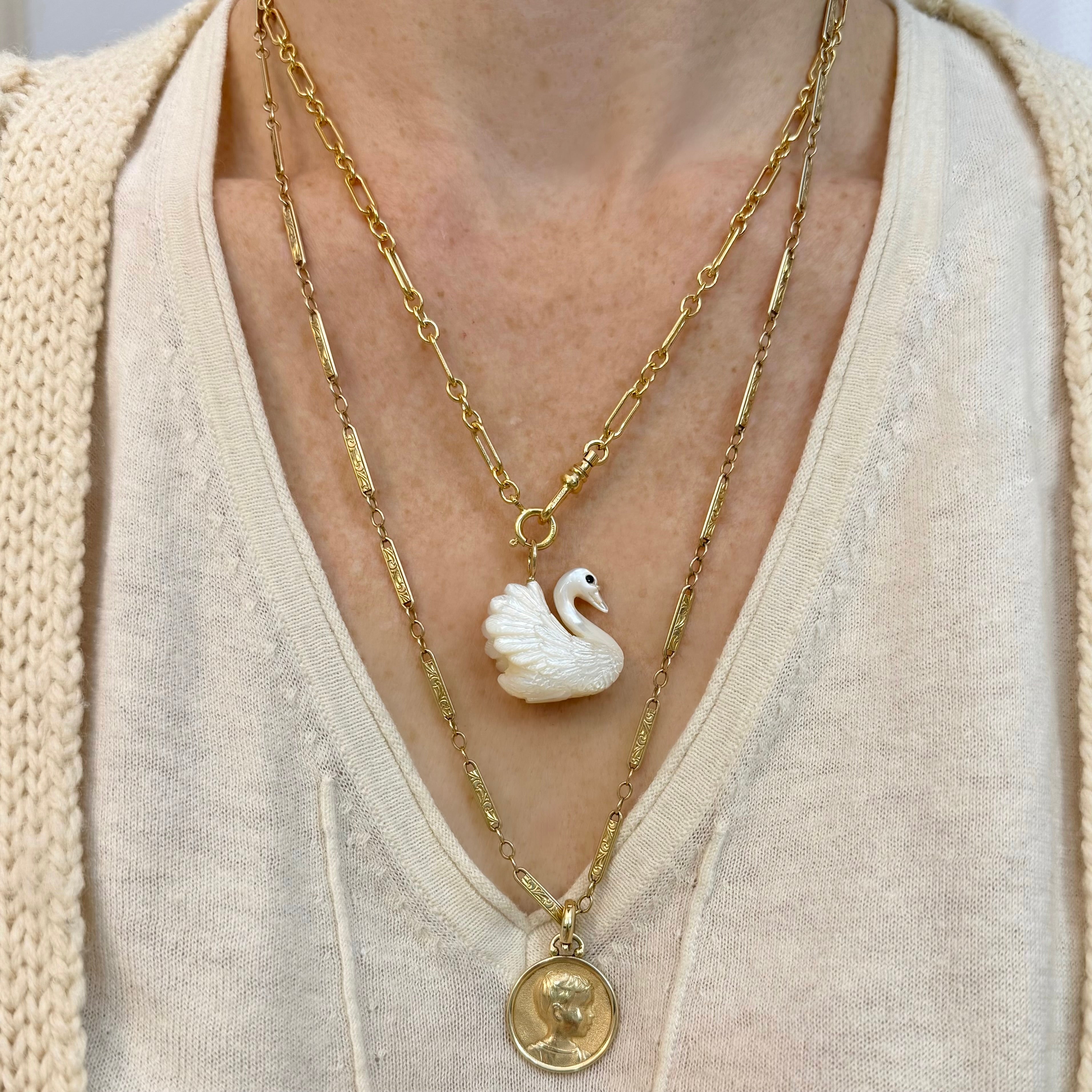 Gold necklace with a white mother of pearl swan charm pendant worn by a person in a beige outfit.