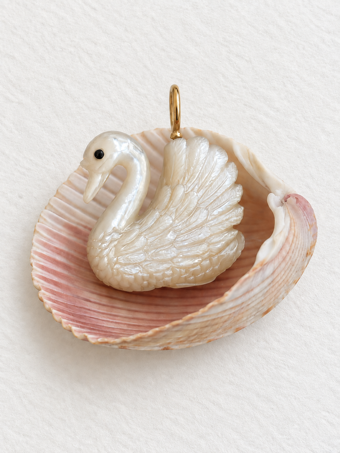 A hand Carved Mother of Pearl realistic swan charm is nestled in a pink scallop shell against a white paper background. Swan charm is designed by Maura Green