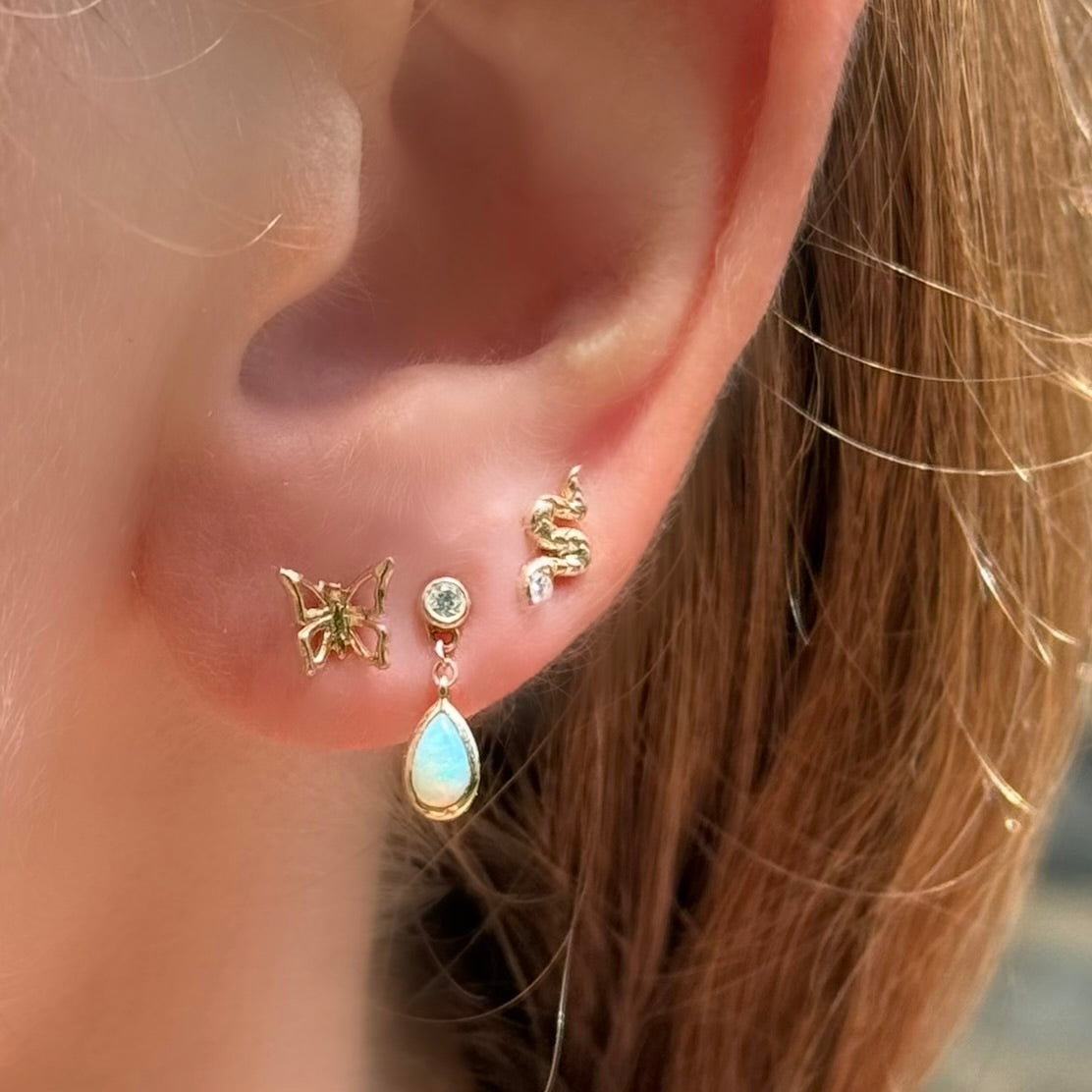 Close-up of an ear with three gold earrings: a butterfly, a teardrop-shaped earring with a gemstone, and another gold earring.