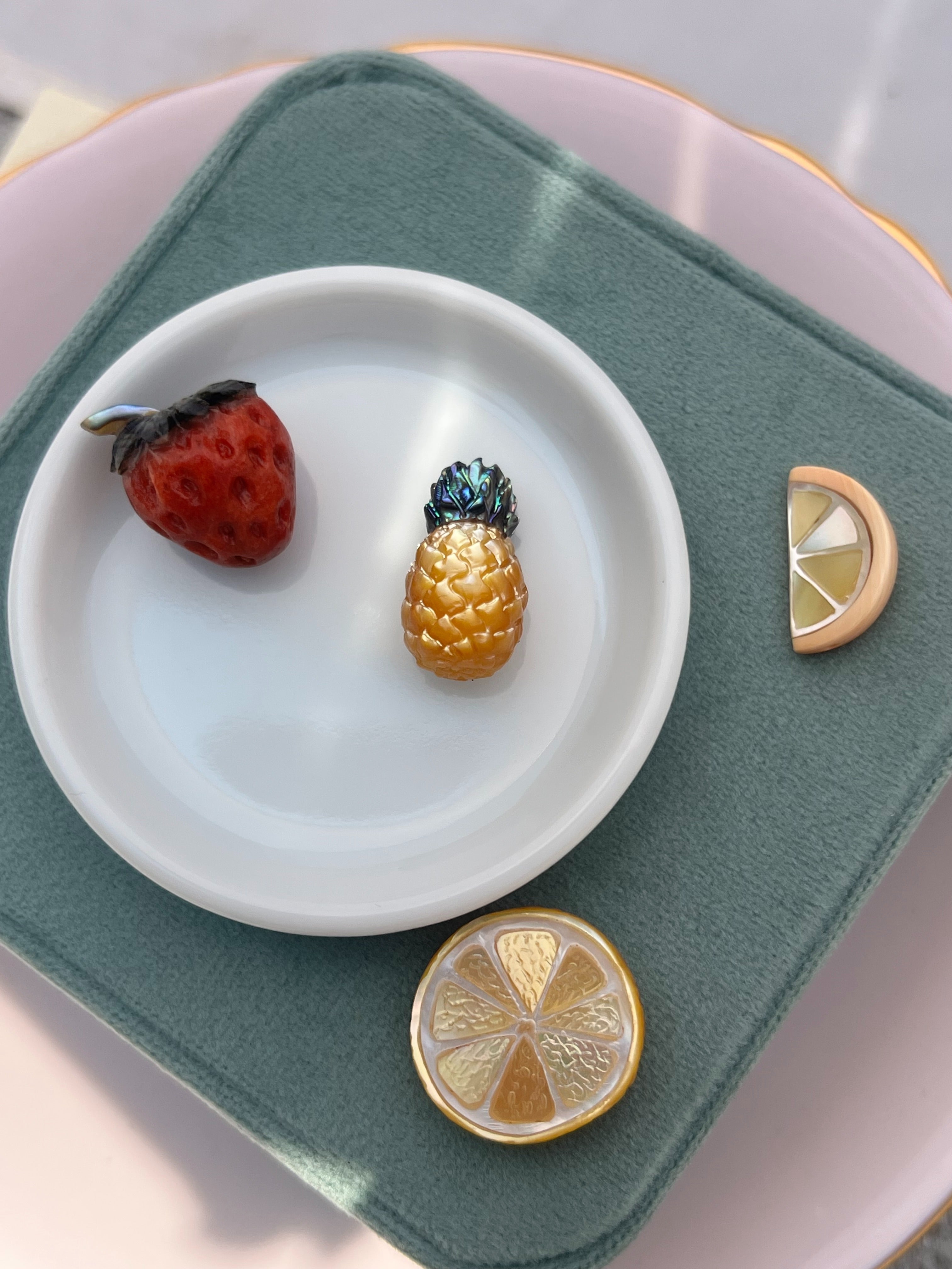 Jewelry charms shaped like a strawberry, pineapple, lemon, and lime on a white dish with a green cloth background.