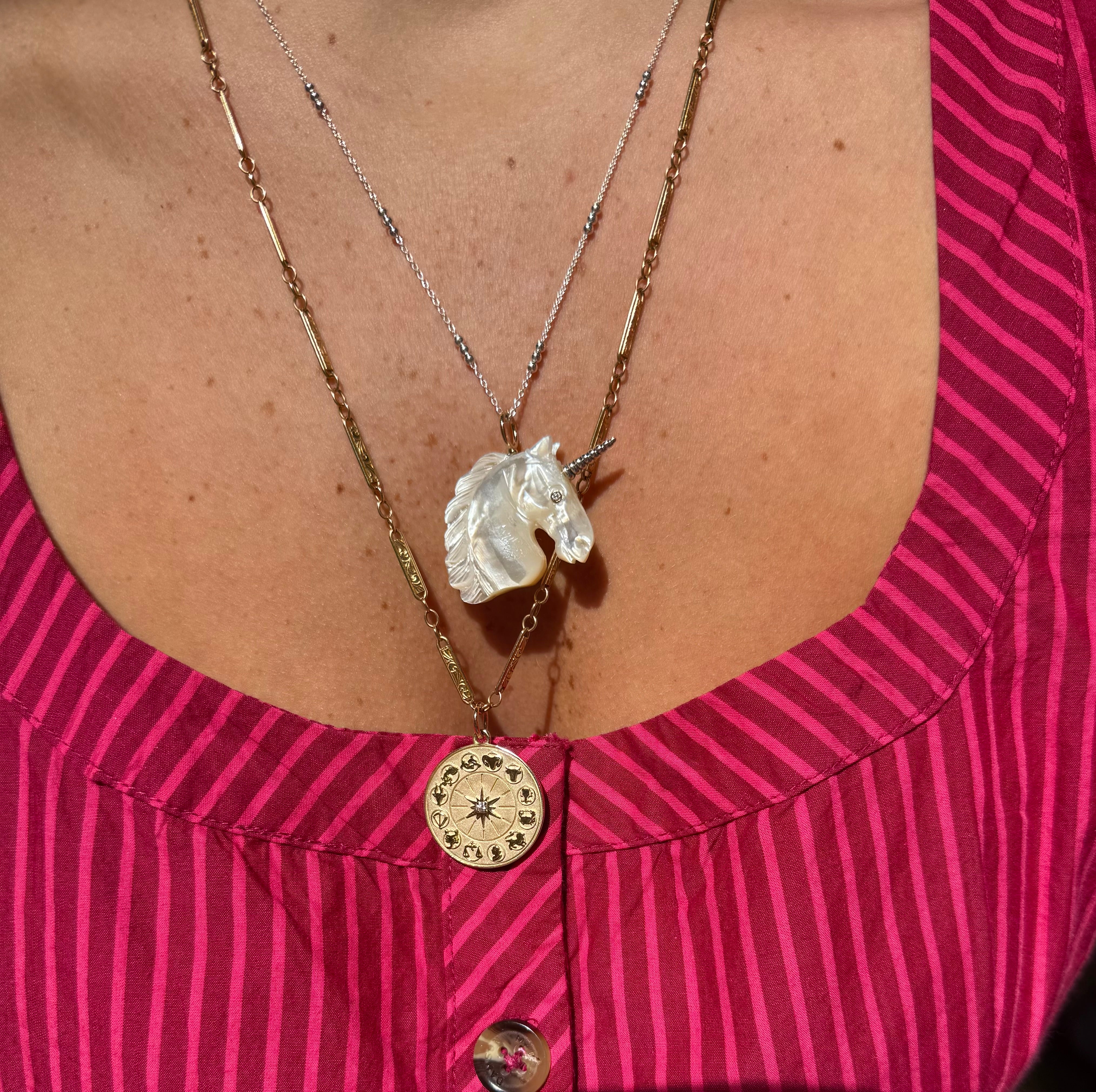 Necklaces with a white unicorn pendant and a Maura Green gold zodiac wheel charm worn by a person in a red striped shirt.