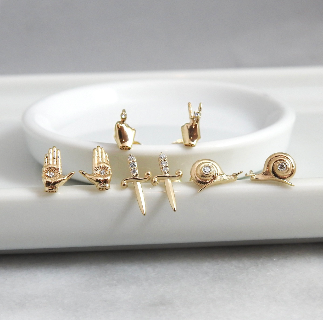 Close up shot of mini yellow gold stud earrings with diamond accents including a middle finger stud, a peace sign hand stud, a pair of hamsa studs, a pair of dagger studs, and a pair of snail studs on a white porcelain background.