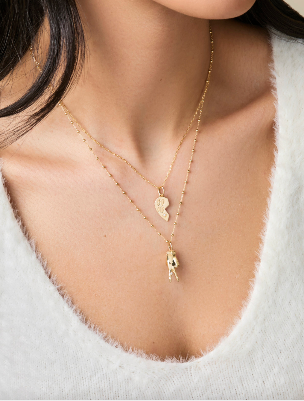 A cropped shot of a woman in a white fuzzy scooped neck shirt wearing two delicate gold necklaces by Maura Green Jewelry including a soul sisters charm and a peace sign ladyfinger charm.