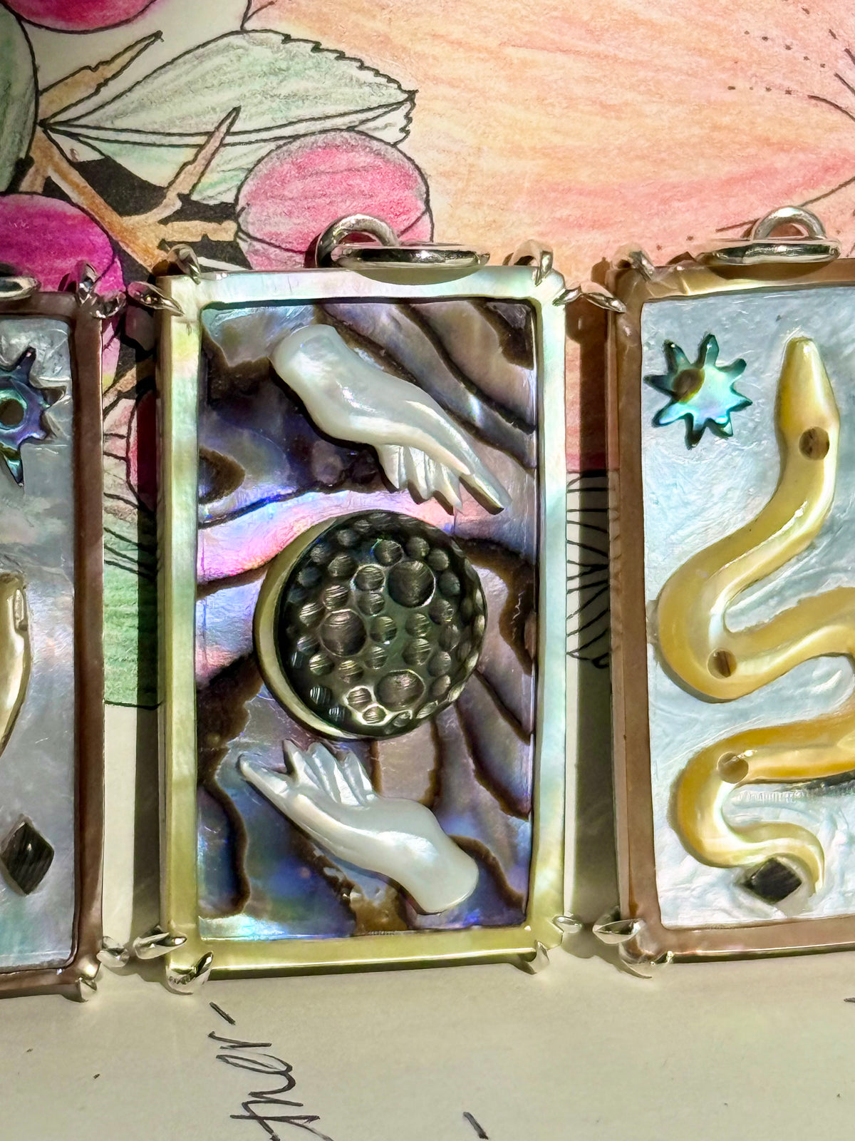 A hand carved abalone and mother of pearl tarot card charm featuring a realistic looking brown mother of pearl moon surrounded by a purple iridescent abalone sky and framed by white mother of pearl hands before a colorful crayon background. Designed by Maura Green